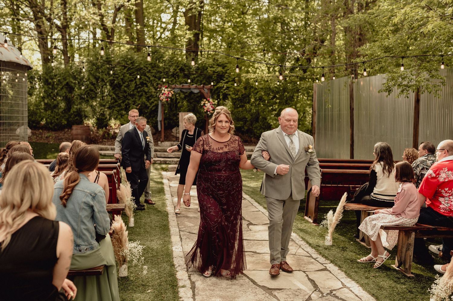 Wedding guests gather outdoors in a wooded setting during a rustic outdoor ceremony.
