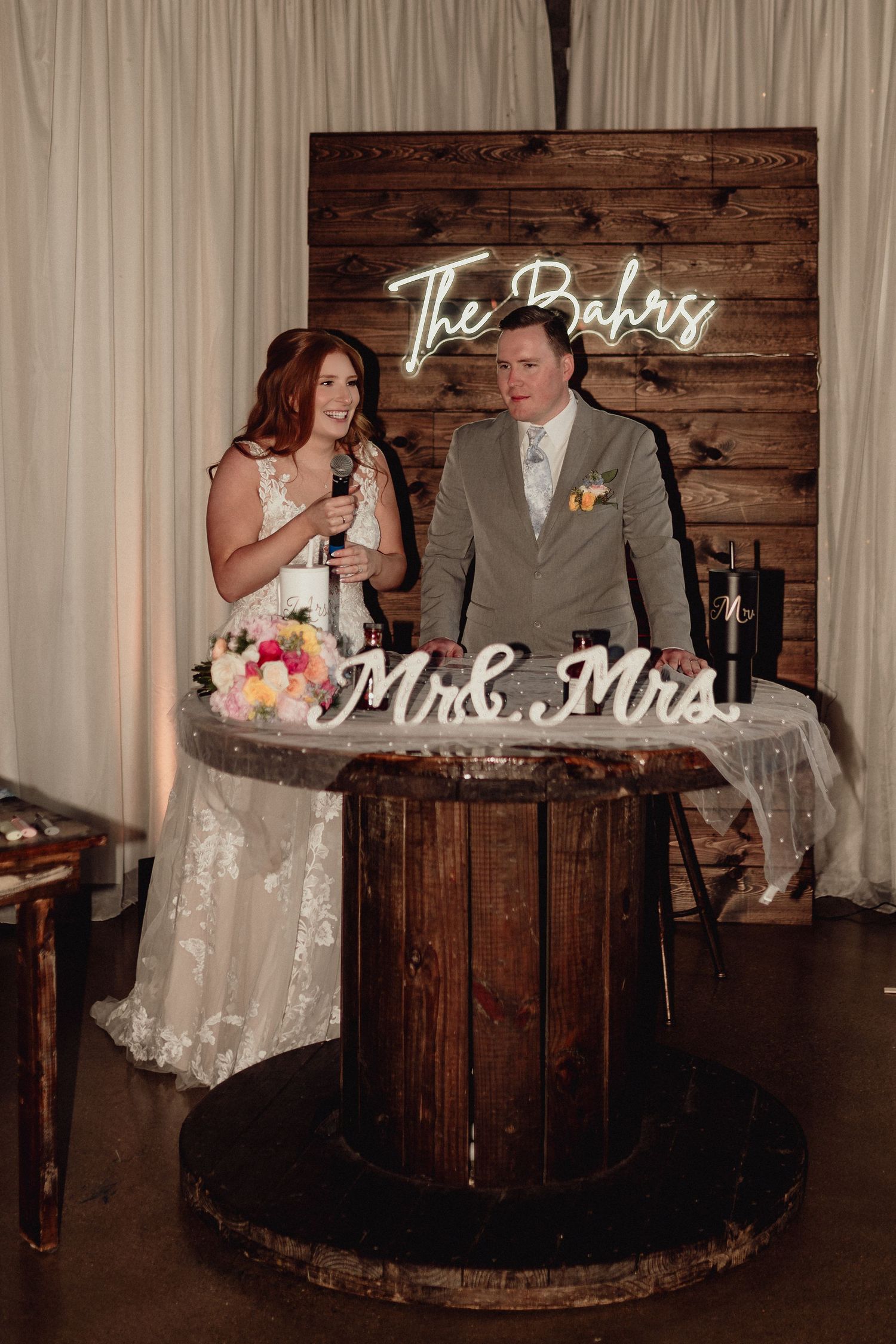 Rustic wedding scene with couple sharing cake at wooden barrel table with Mrs and Mrs neon sign glowing in background.