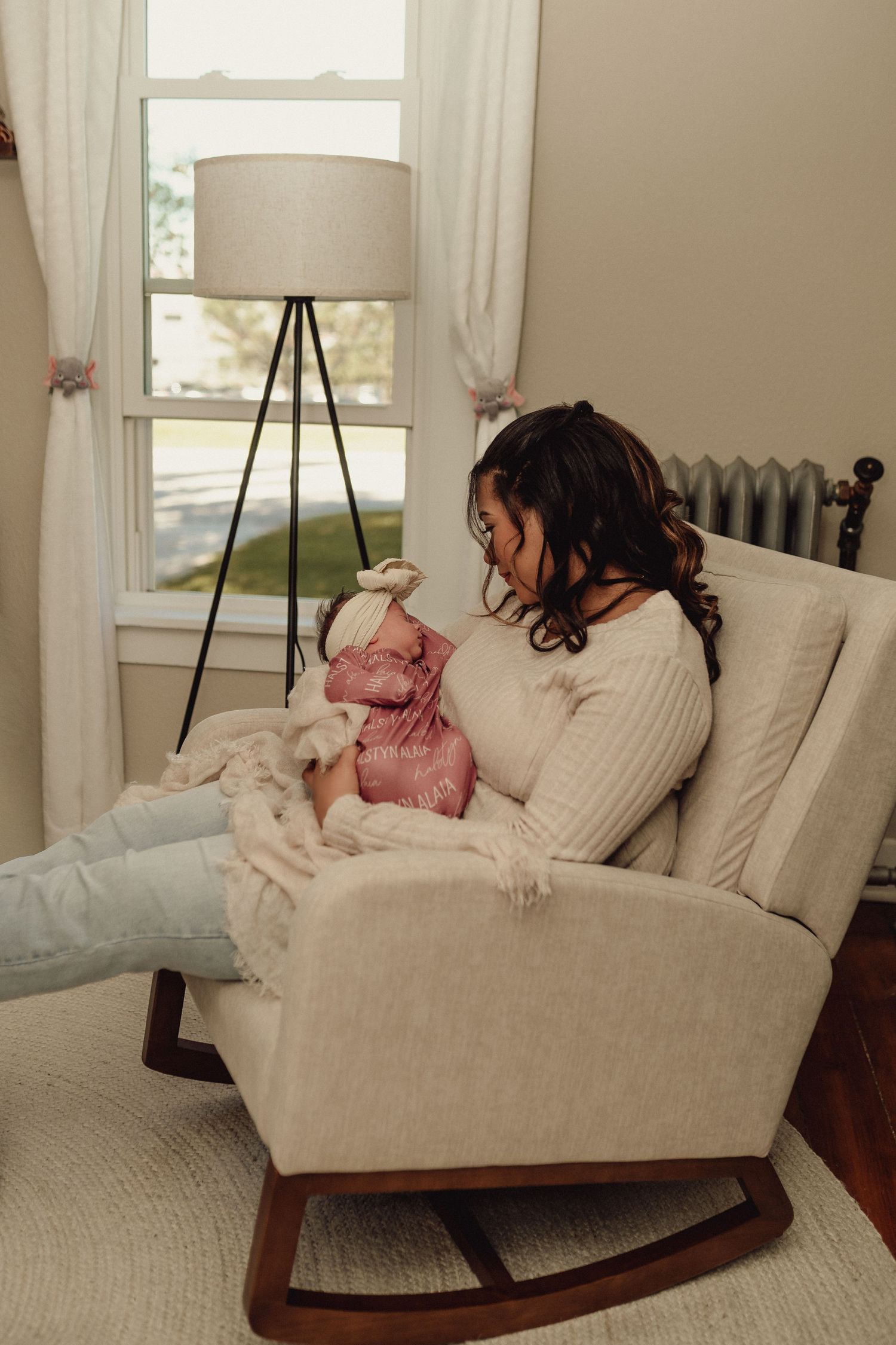 A comfortable beige rocking chair sits in a bright nursery room with white curtains.