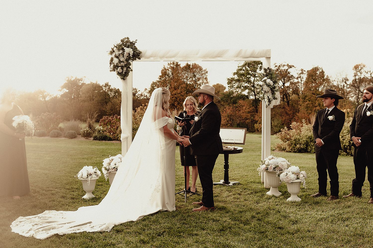 Outdoor wedding ceremony with guests gathered on a grassy field at sunset.