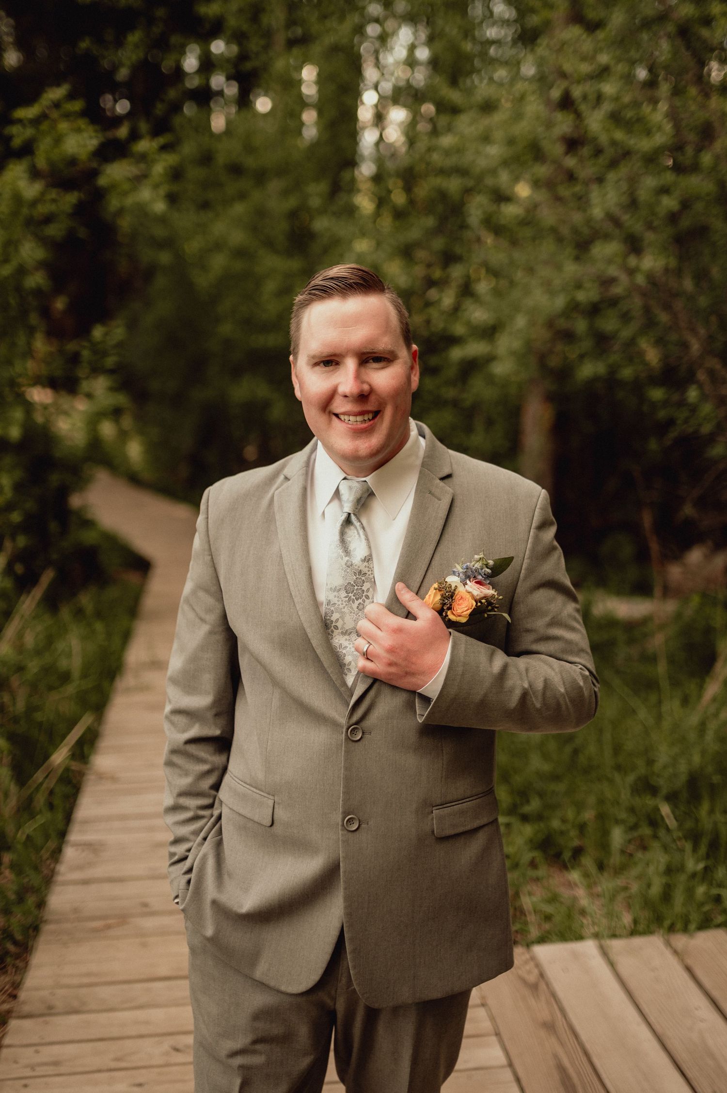 A person in a light gray suit and tie stands on a wooden boardwalk path surrounded by lush green foliage.