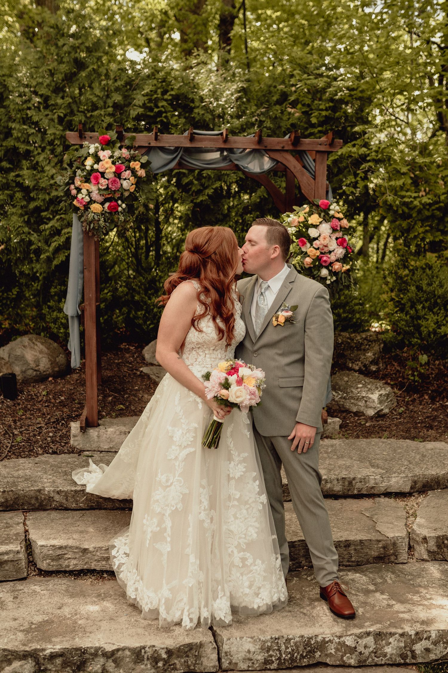 A romantic wedding couple shares a tender kiss in front of a rustic stone wall decorated with pink floral arrangements.