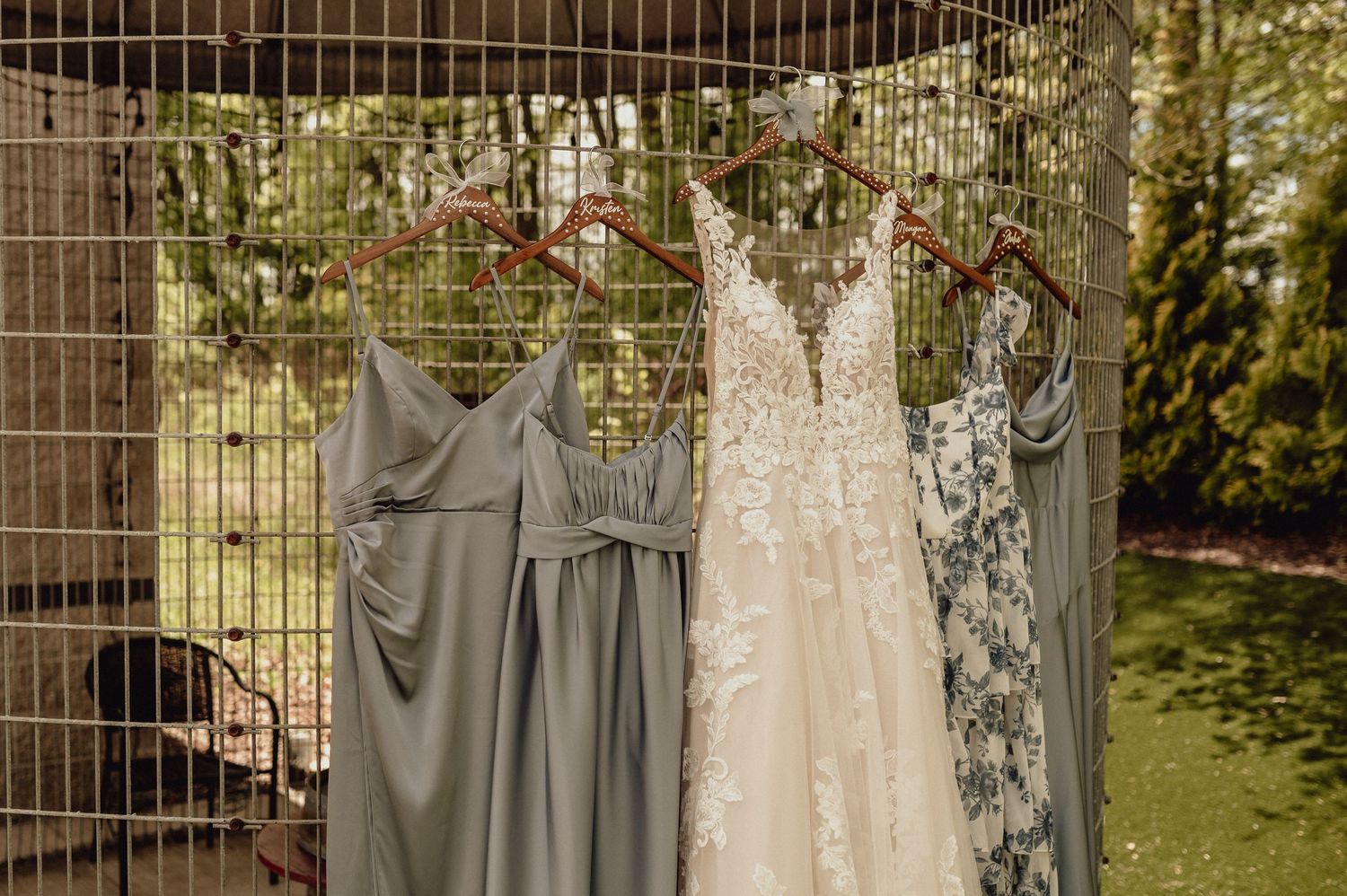 Wedding dresses and gray bridesmaid gowns hang on a wooden wall in natural outdoor lighting.