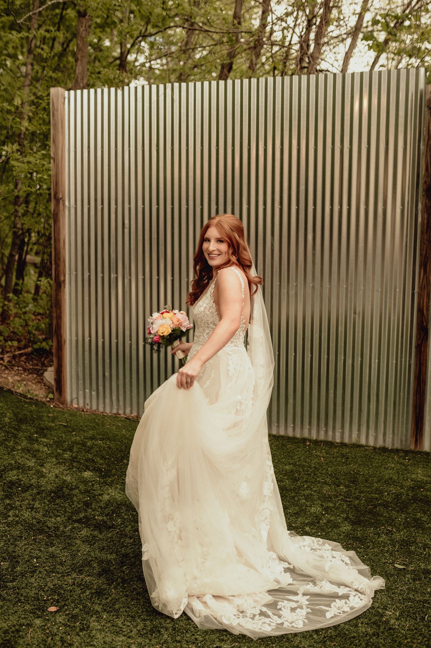 A bride in a flowing lace wedding gown poses gracefully against a rustic metal wall backdrop in an outdoor garden setting.