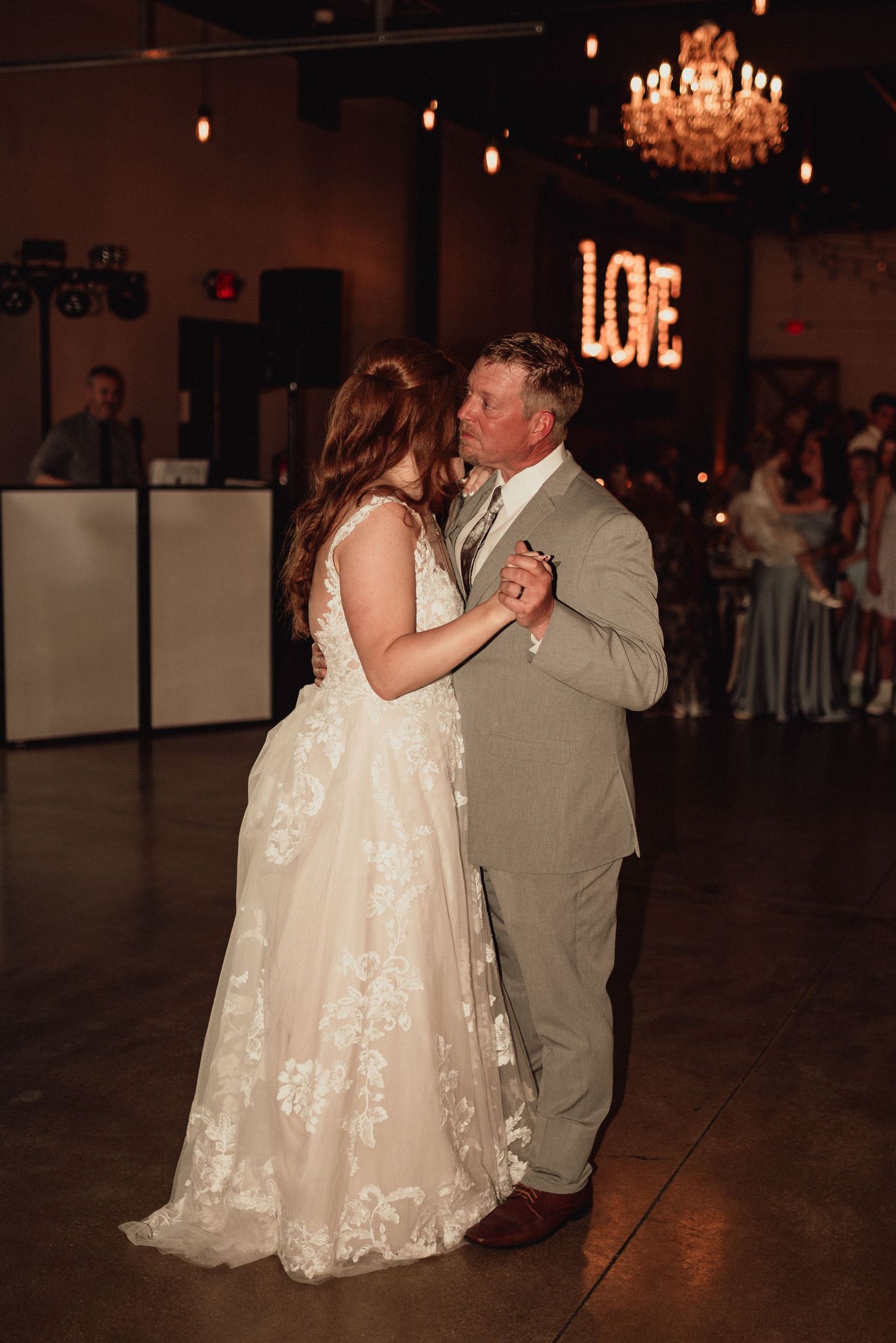 A couple shares their first dance at a wedding reception under a crystal chandelier with illuminated 'LOVE' sign on the wall.