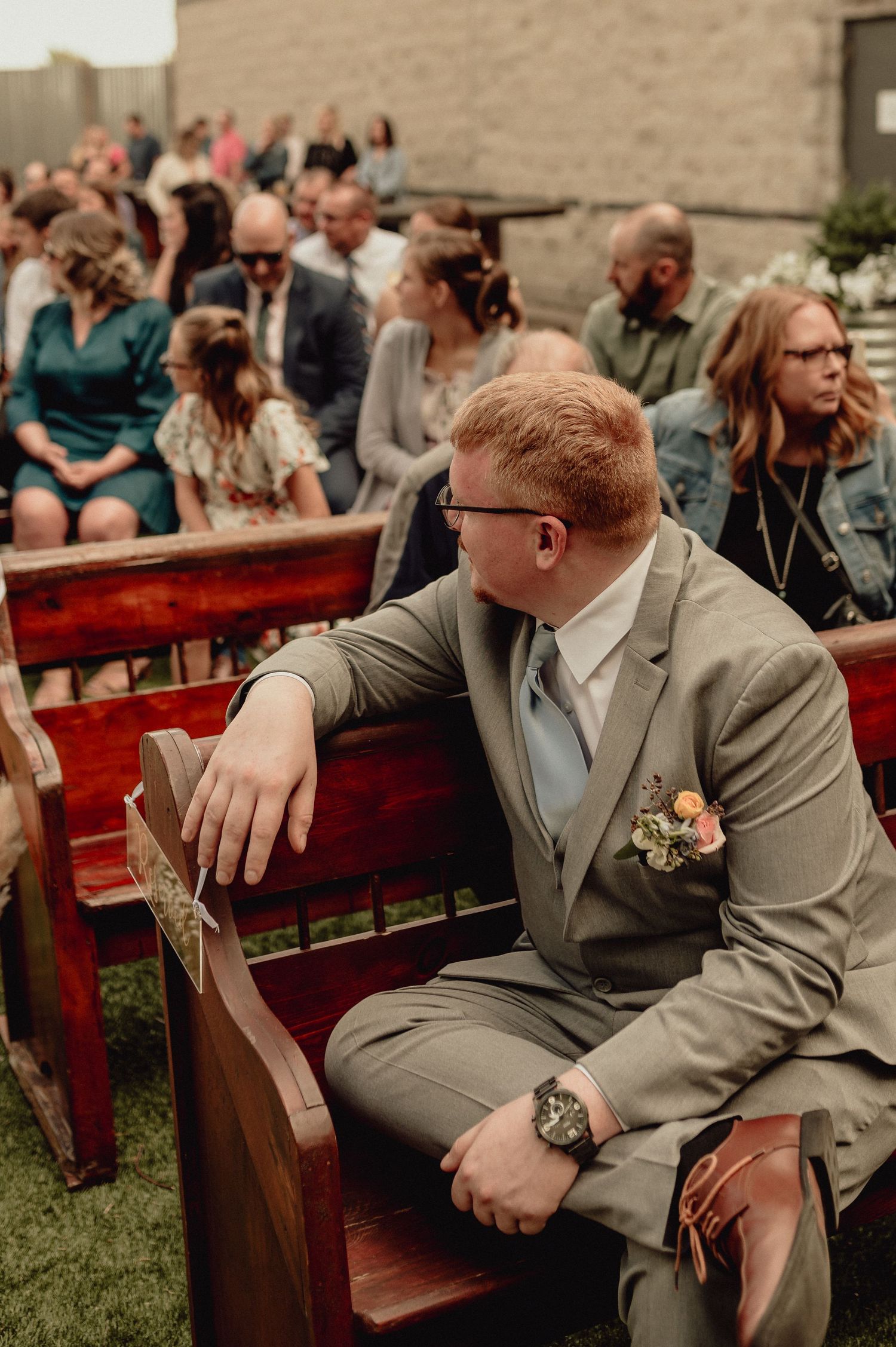 Wedding guests sit on rustic wooden benches at an outdoor ceremony with a person in a gray suit in the foreground.
