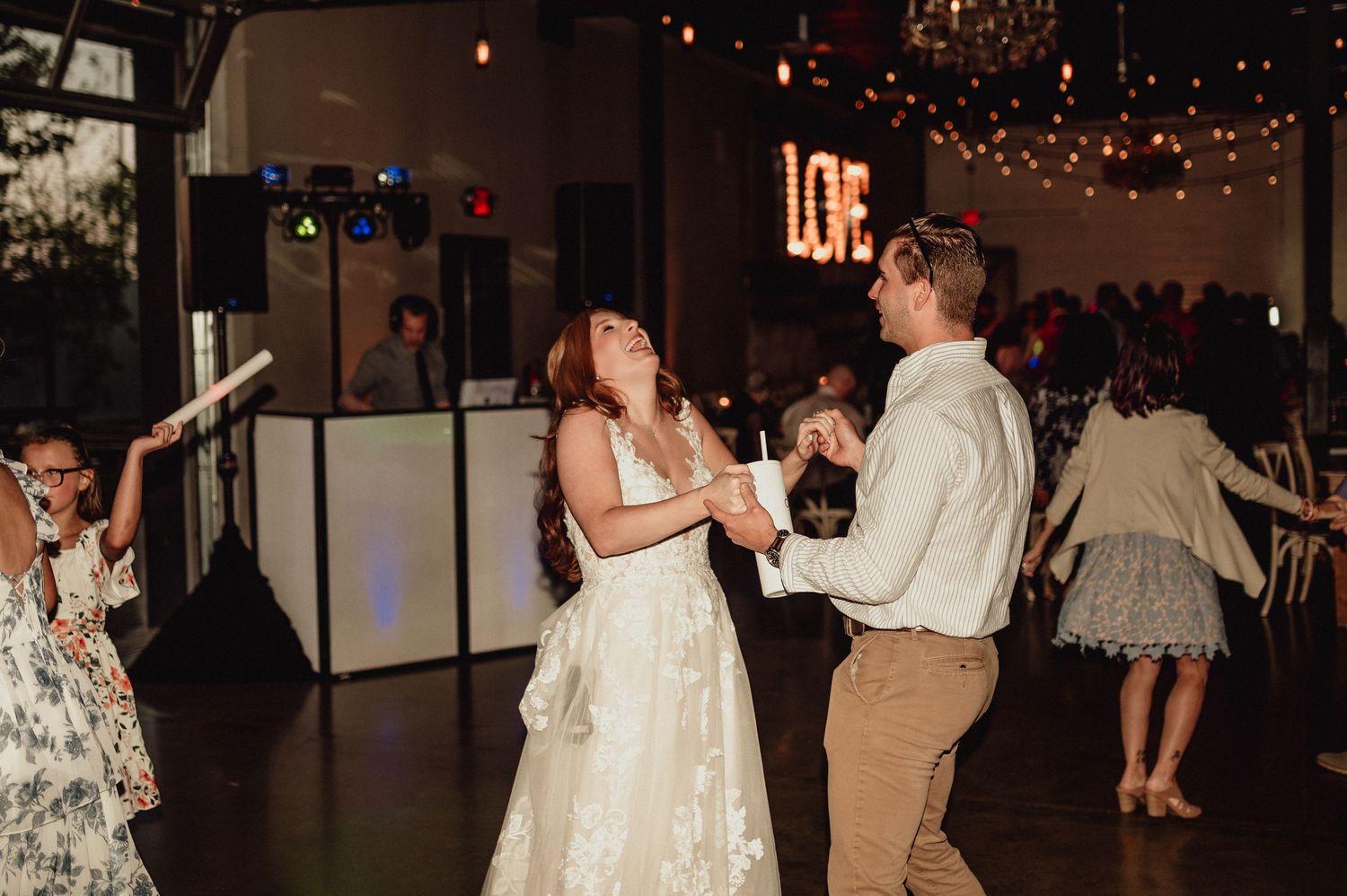 Wedding reception guests dance and celebrate on a dimly lit dance floor decorated with string lights.
