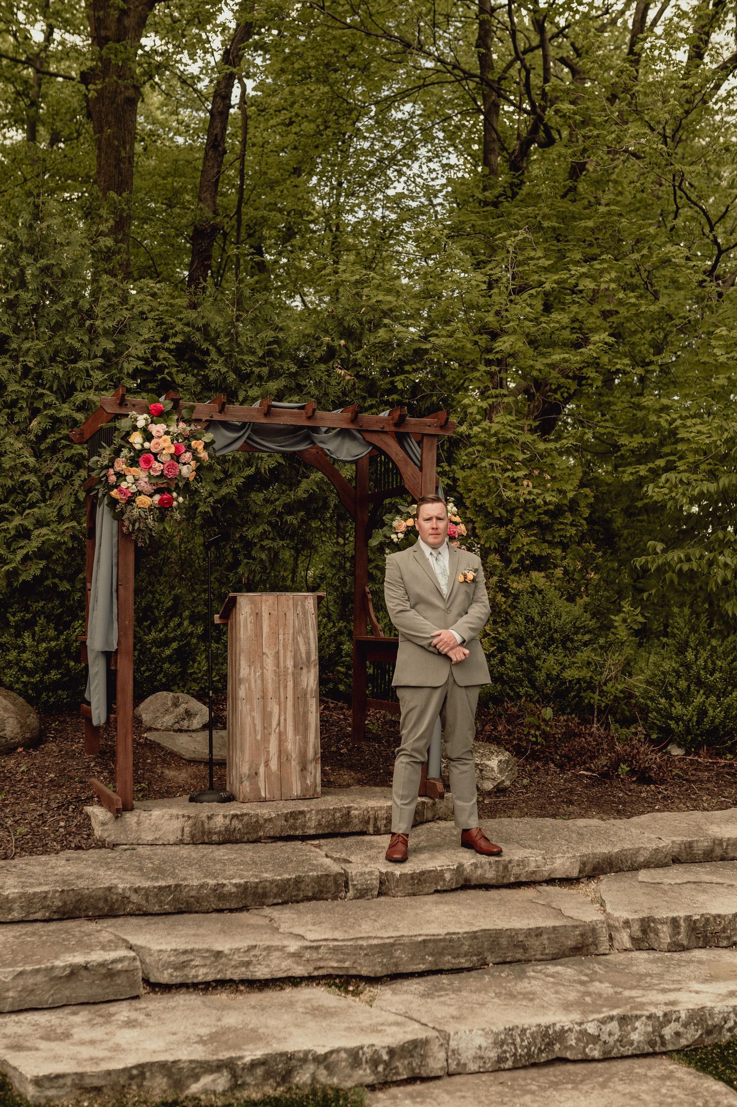 A figure sits on a rustic stone wall in a garden setting with lush greenery and floral decorations.