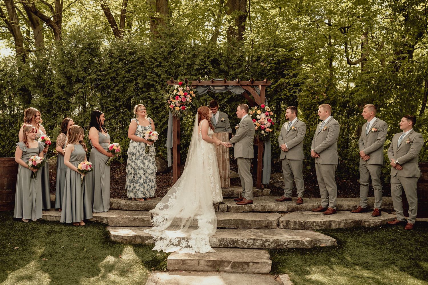 Wedding party stands in a lush garden setting with stone steps and greenery creating an elegant outdoor backdrop.