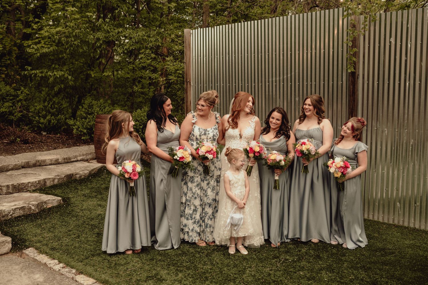 A wedding party in gray dresses poses outdoors against a corrugated metal wall on a sunny day.
