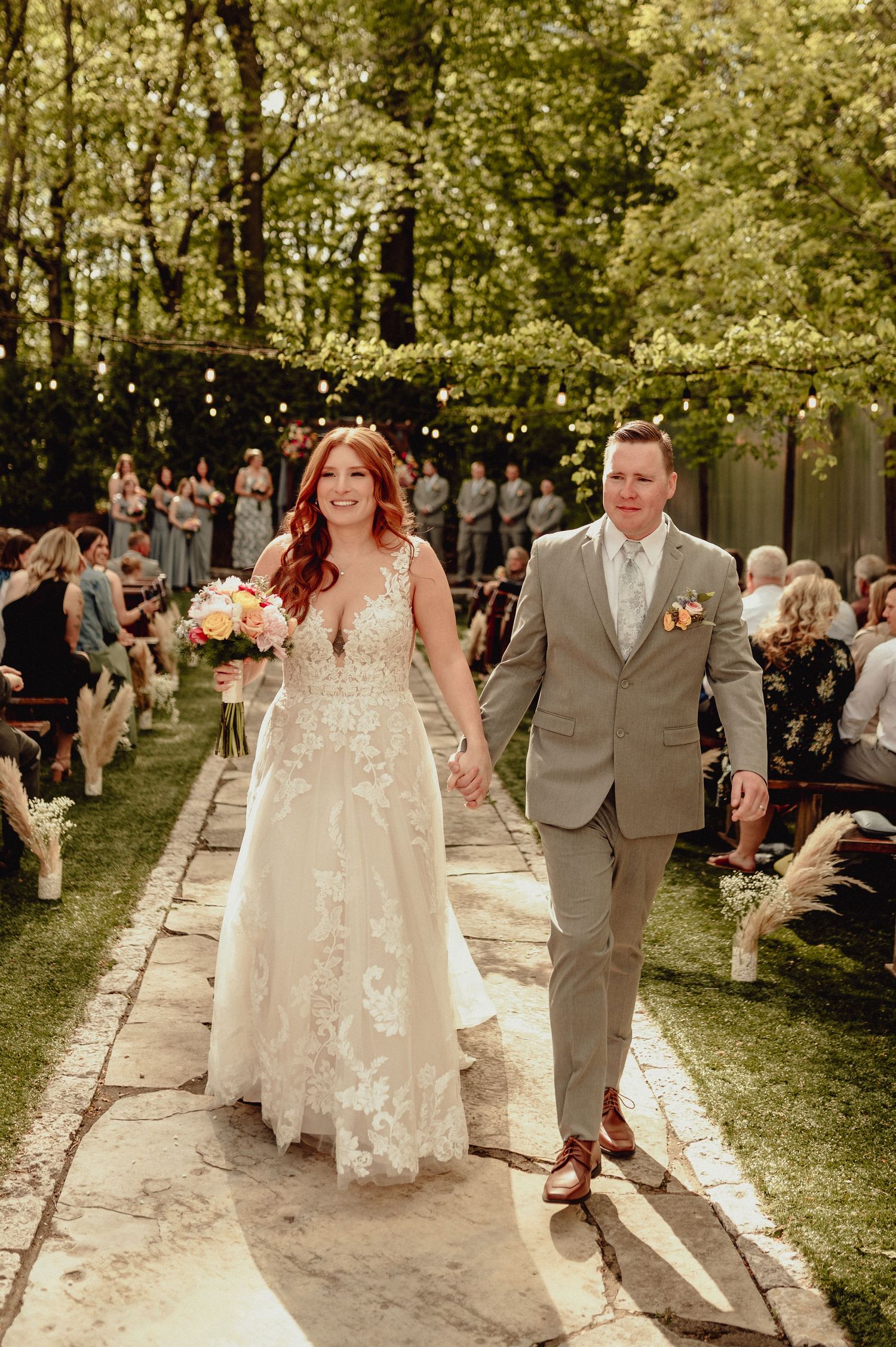 A wedding ceremony takes place on a stone path surrounded by lush trees and greenery in a beautiful outdoor setting.