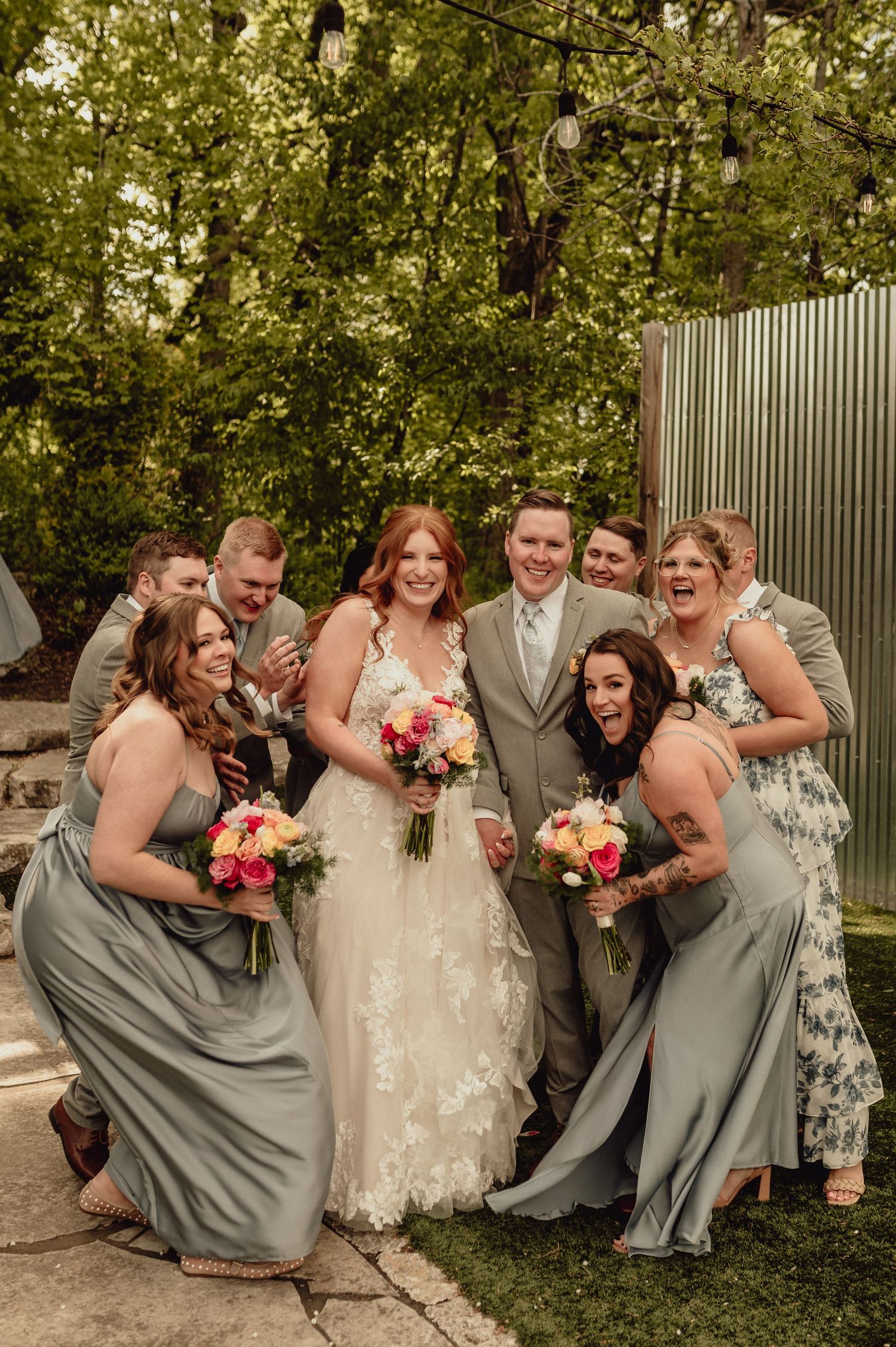 Wedding party laughs together while posing outdoors in gray and floral attire with pink bouquets.