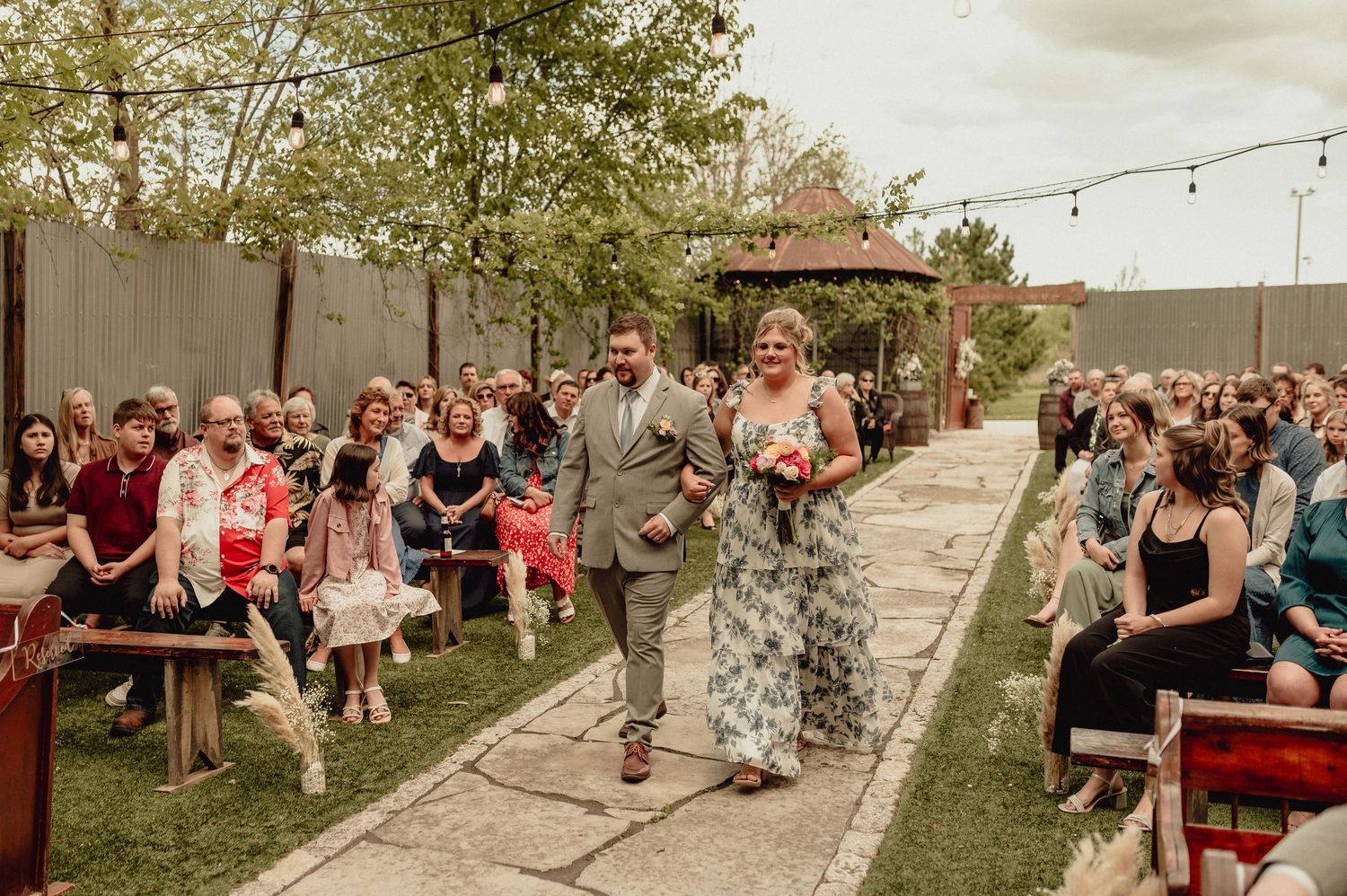 Outdoor wedding guests gather on a grassy lawn under string lights during a sunset celebration.