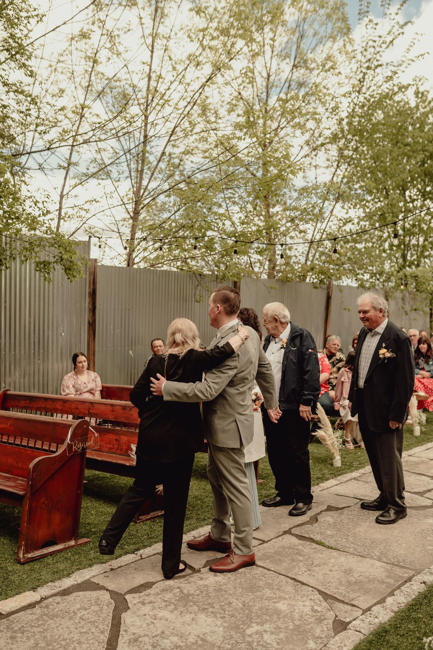Wedding guests dance and celebrate together in a line formation outdoors on a sunny day.