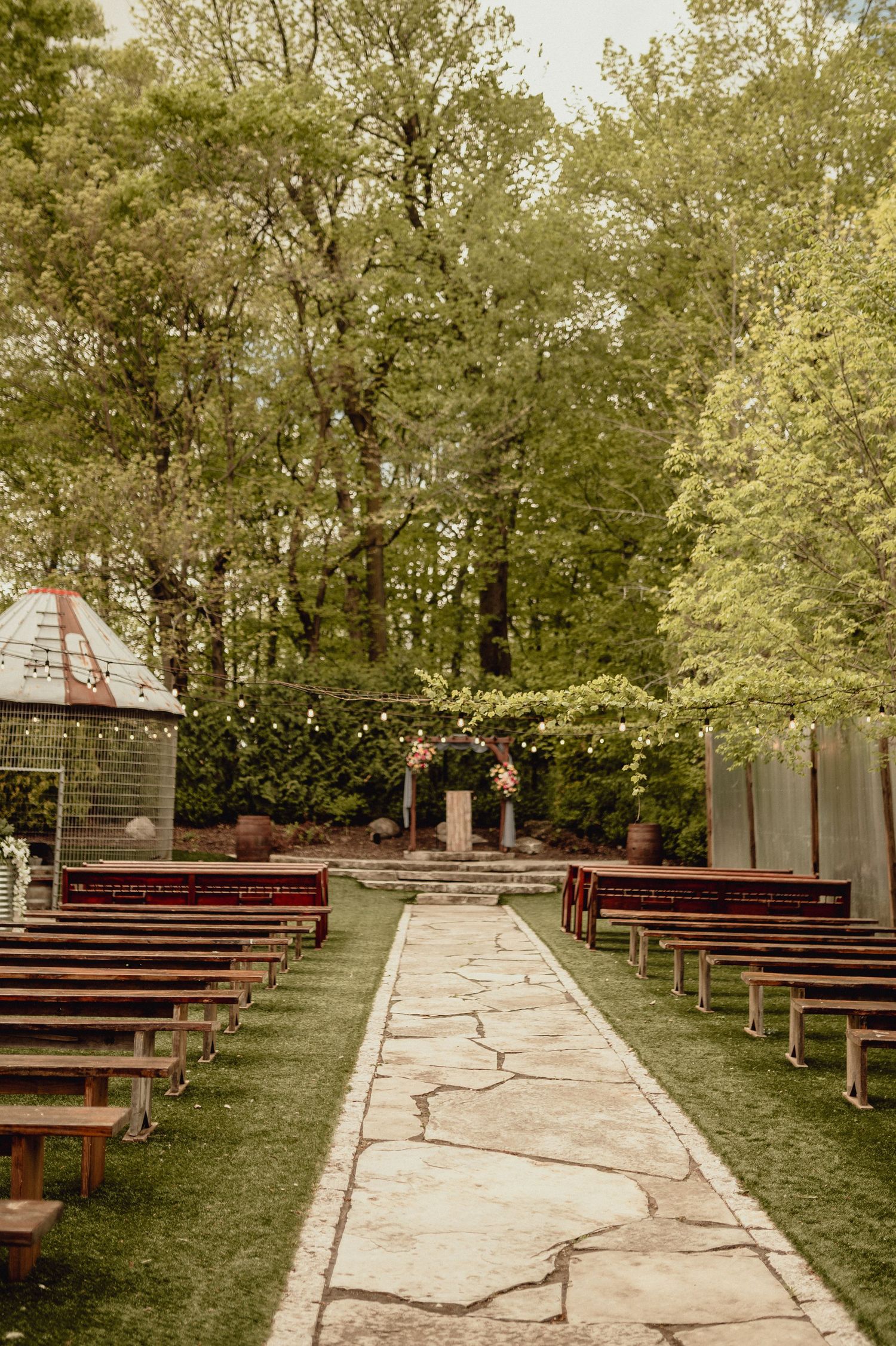 A stone pathway leads between wooden benches to an outdoor wedding ceremony setup in a wooded garden setting.
