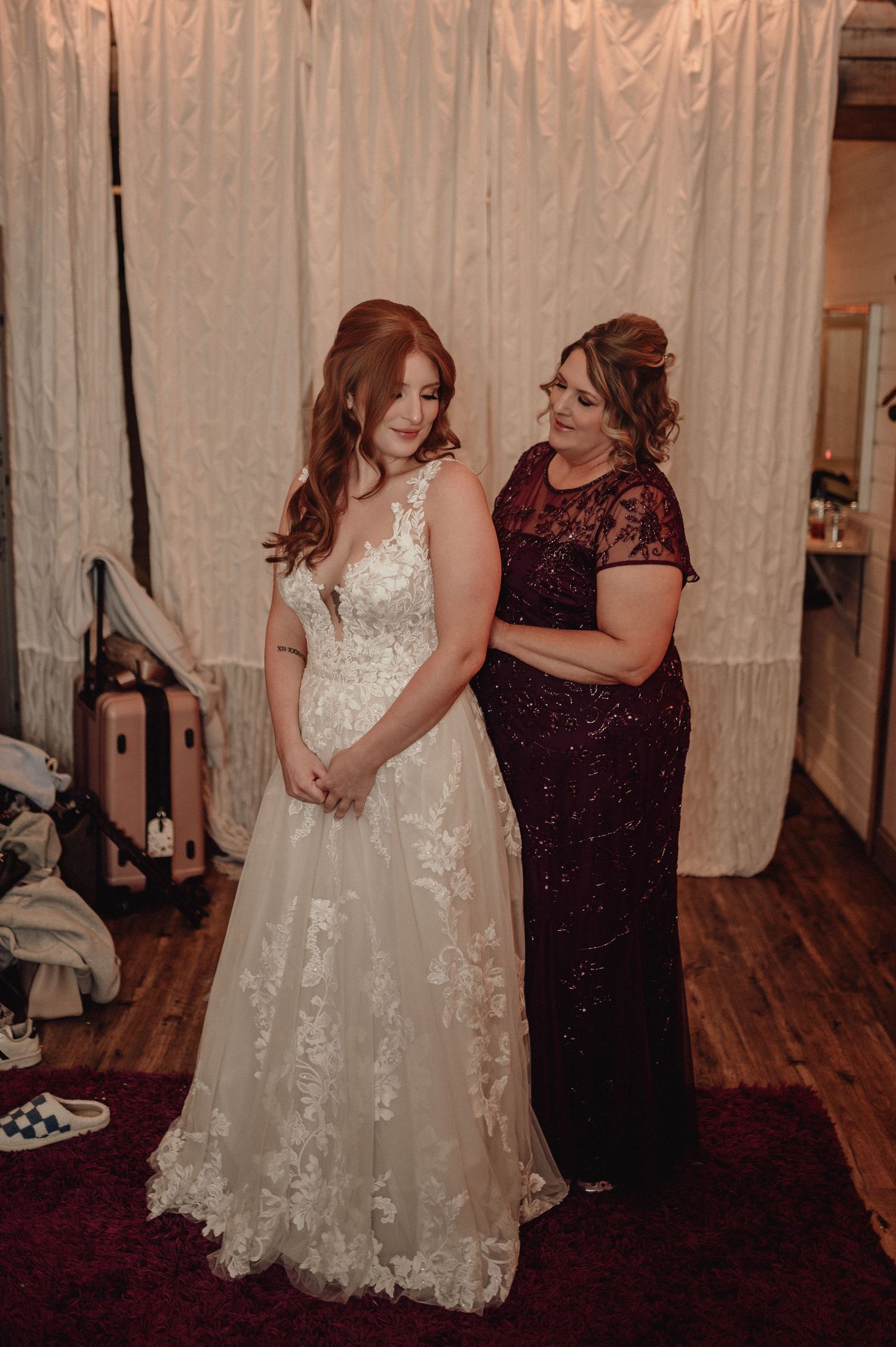 A bride in a white lace dress stands next to someone wearing a burgundy gown during a wedding fitting.
