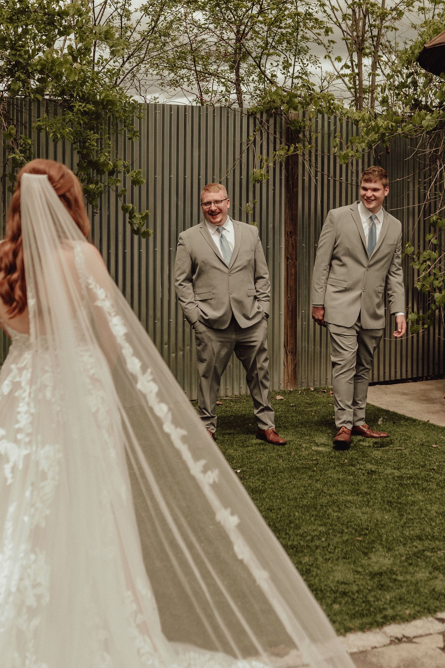 A wedding party in gray suits and a flowing white bridal gown pose in a backyard garden setting.