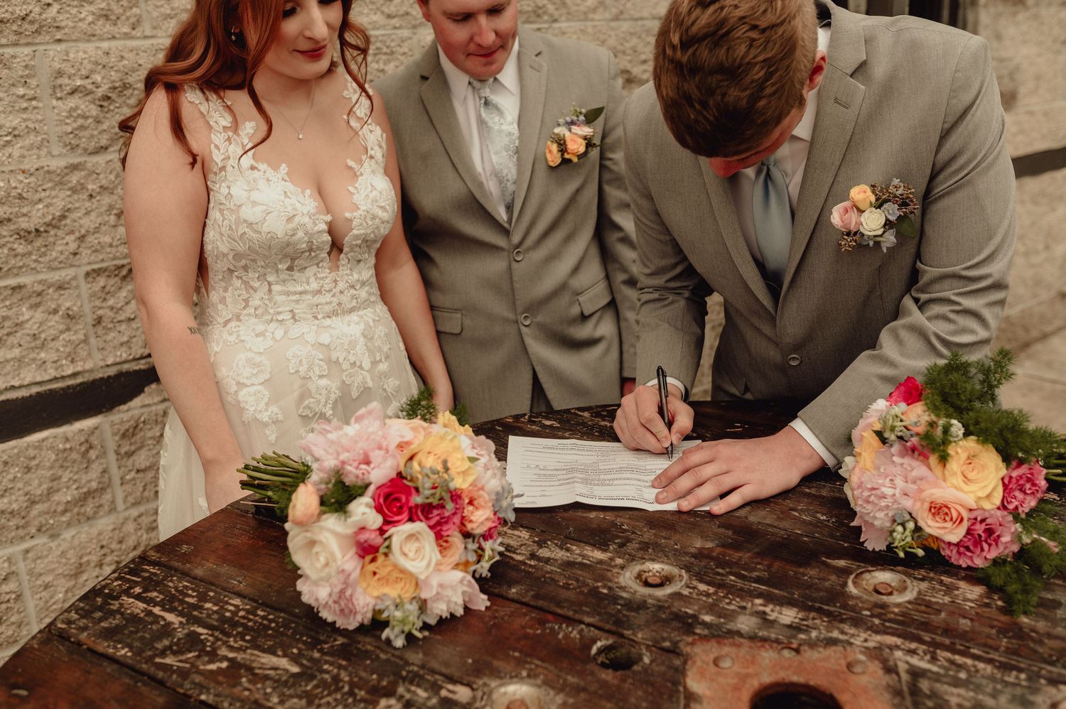 Wedding couple and officiant signing marriage documents on rustic wooden table with pink and white floral bouquets.