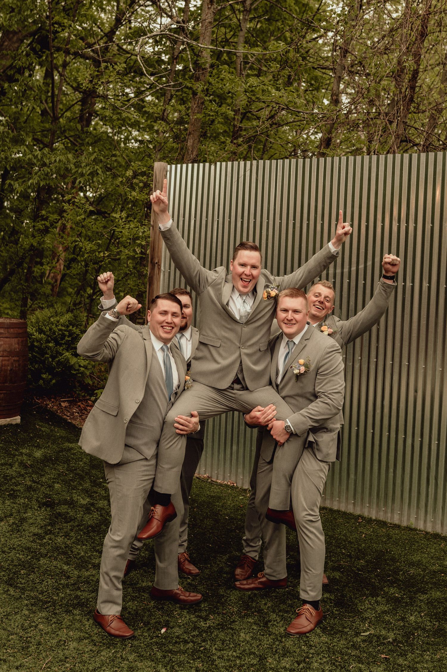 A group of groomsmen in light gray suits pose playfully against a corrugated metal wall in an outdoor garden setting.