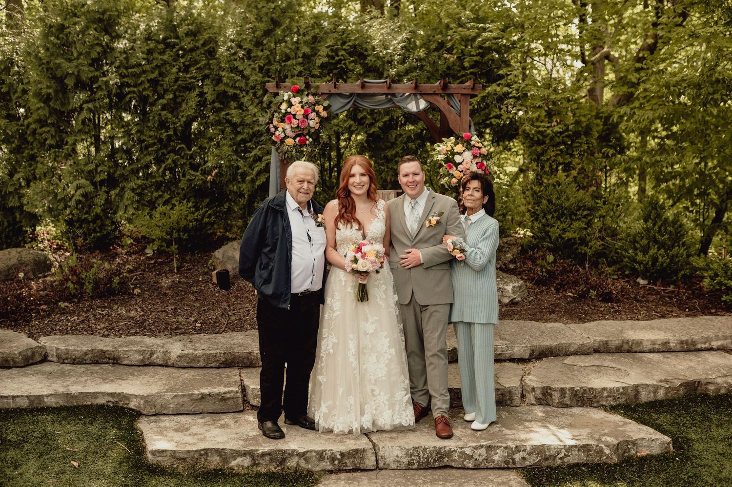 Wedding party poses for formal portraits in a garden setting with stone wall and lush greenery in the background.