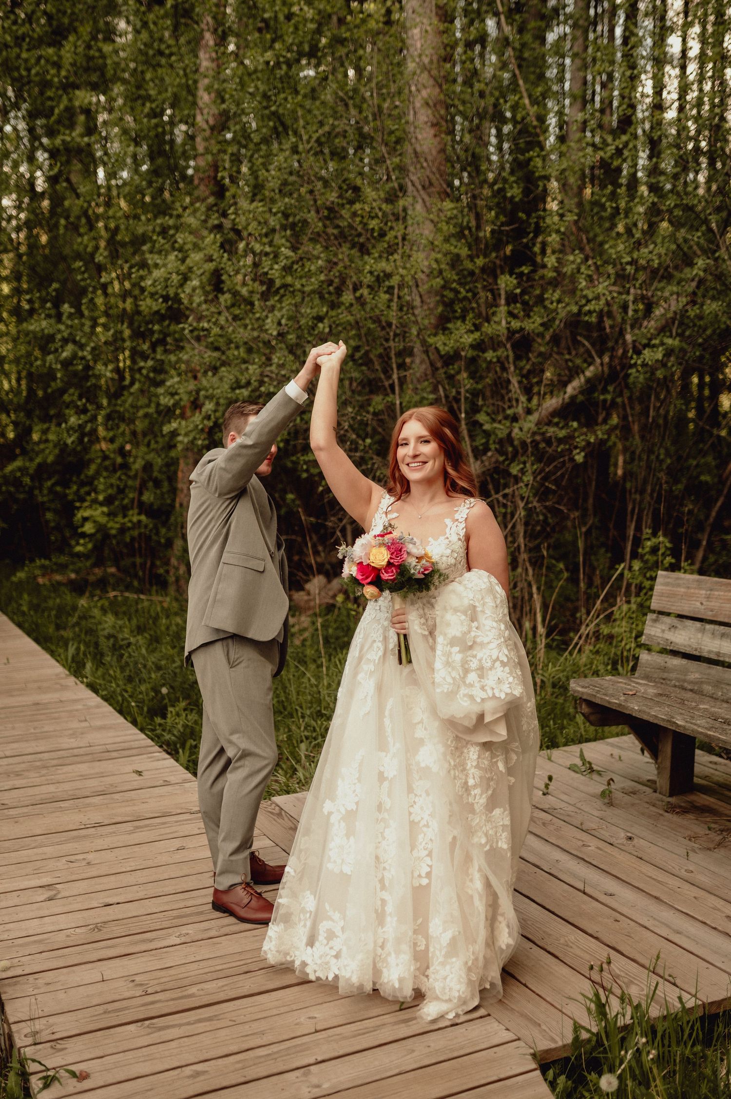 A bride in a lace wedding dress sits on a rustic wooden boardwalk surrounded by lush green forest.