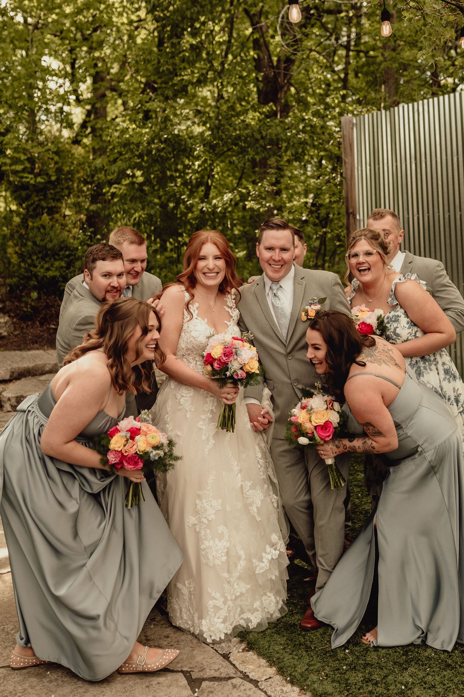 Wedding party in gray attire poses together outdoors while laughing and sharing a joyful candid moment.