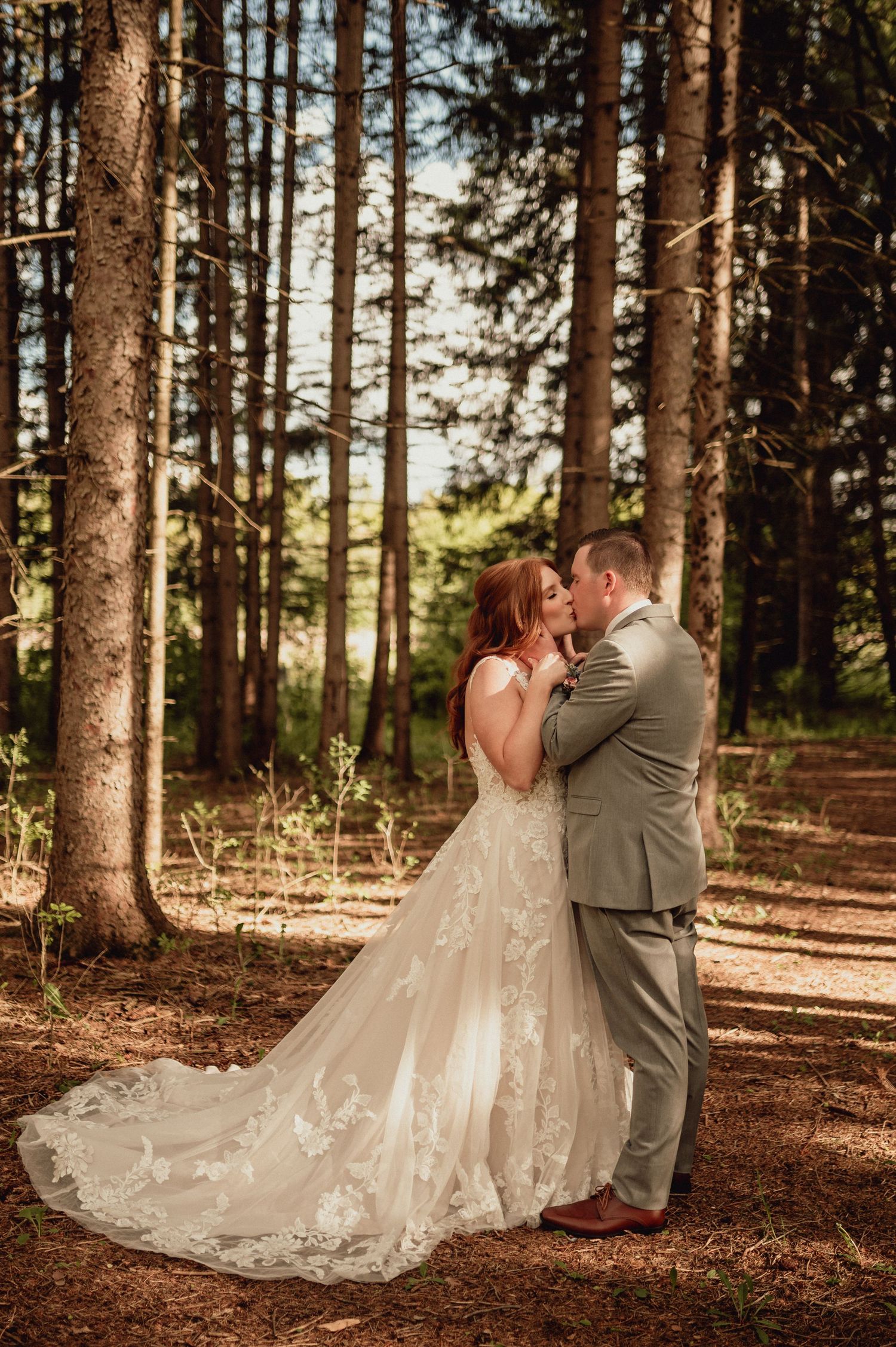 Newlyweds embrace in a wooded setting with sunlight streaming through evergreen trees.