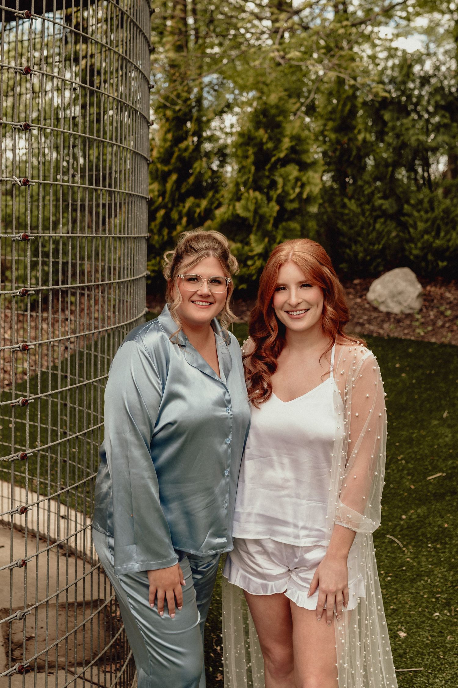 Two people pose together outdoors in casual attire against a natural green background with fence.