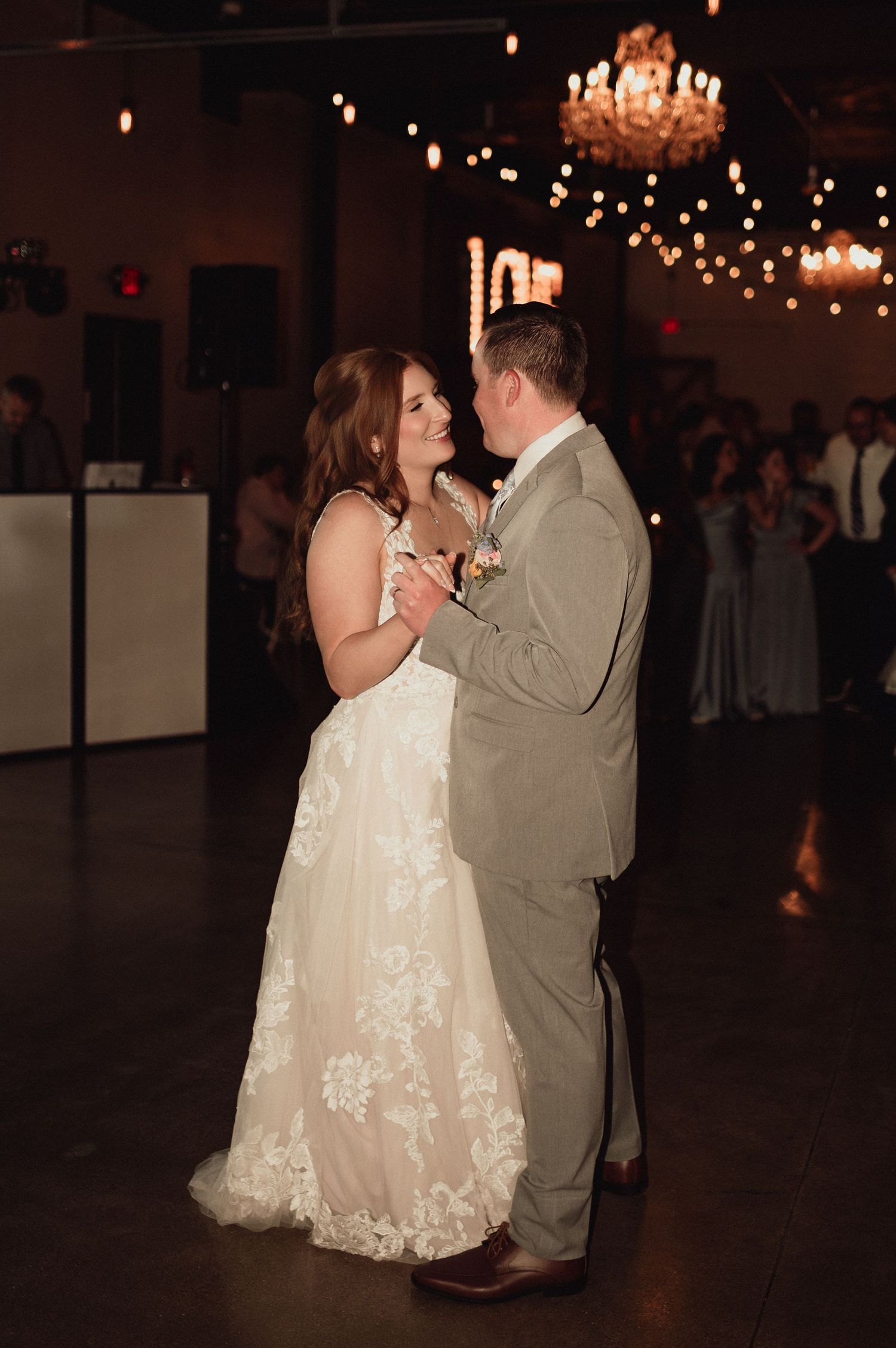 A couple shares their first dance at a wedding reception under a crystal chandelier and string lights.