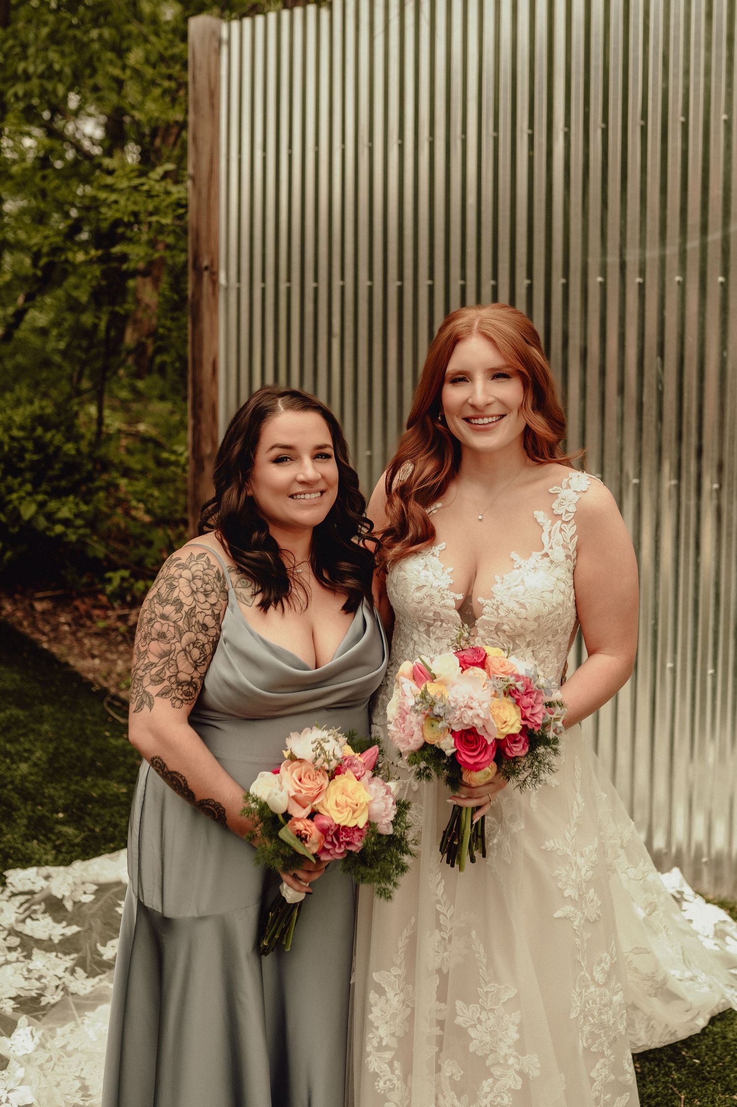 Two people pose together at a wedding, one wearing a gray dress and one in a white lace gown holding floral bouquets.