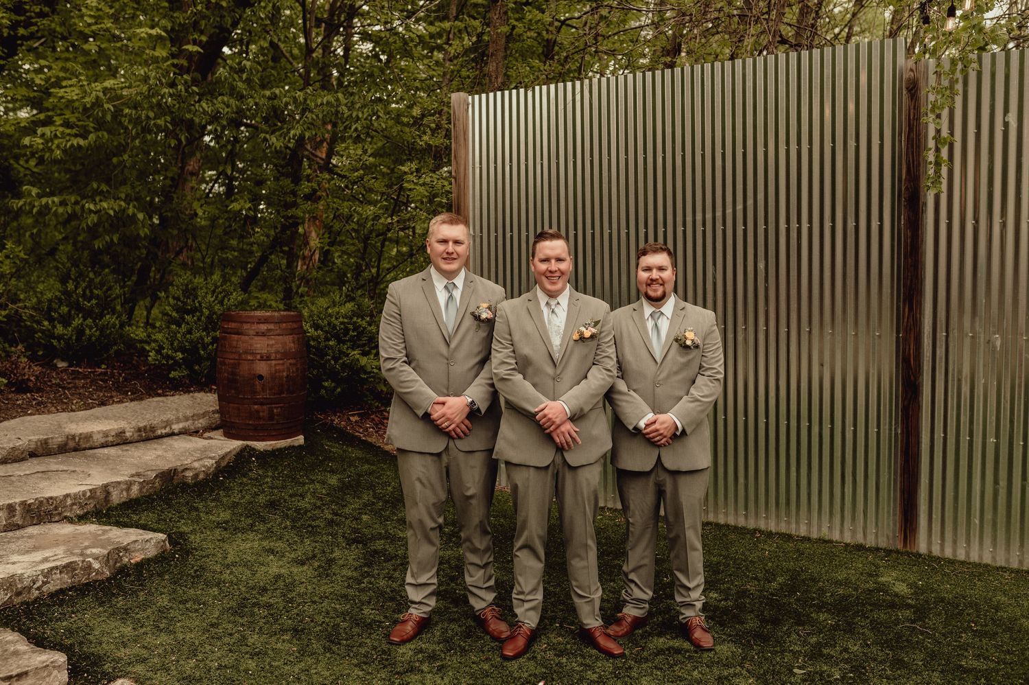 A group in light gray suits poses together in an outdoor garden setting with greenery and a fence in the background.