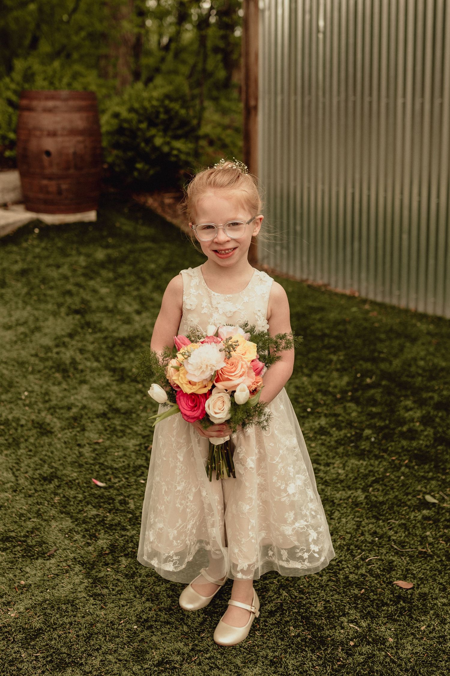A flower girl in a white lace dress holds a peach and pink bouquet while sitting on green grass.