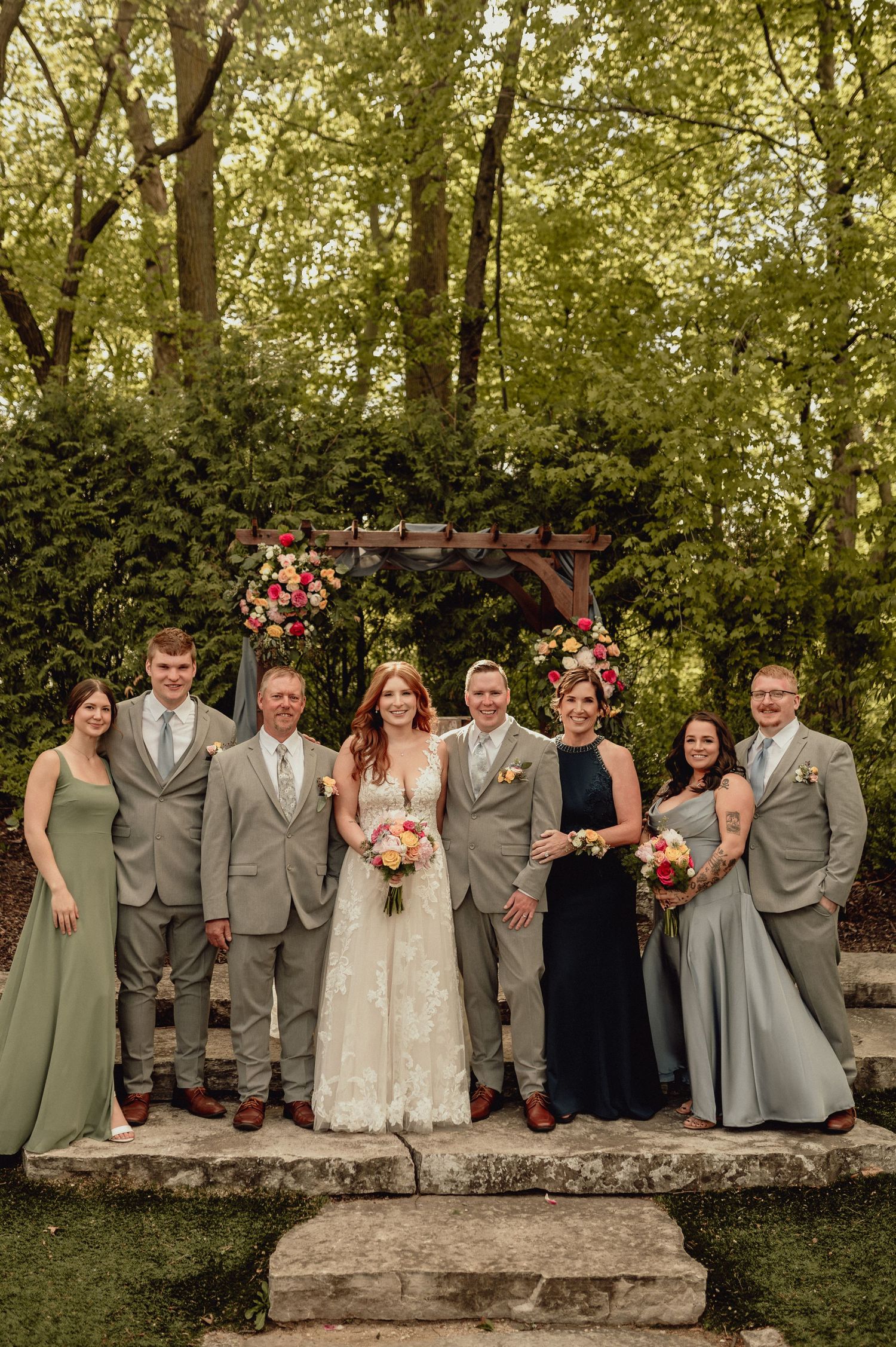 Wedding party in sage green and gray attire poses together on rustic stone wall in outdoor garden setting.