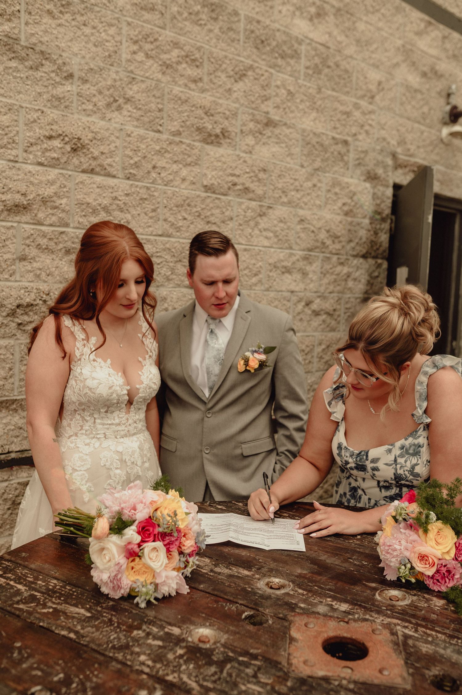 A couple signs their marriage license at a rustic stone wall venue while holding a vibrant pink and peach floral bouquet.