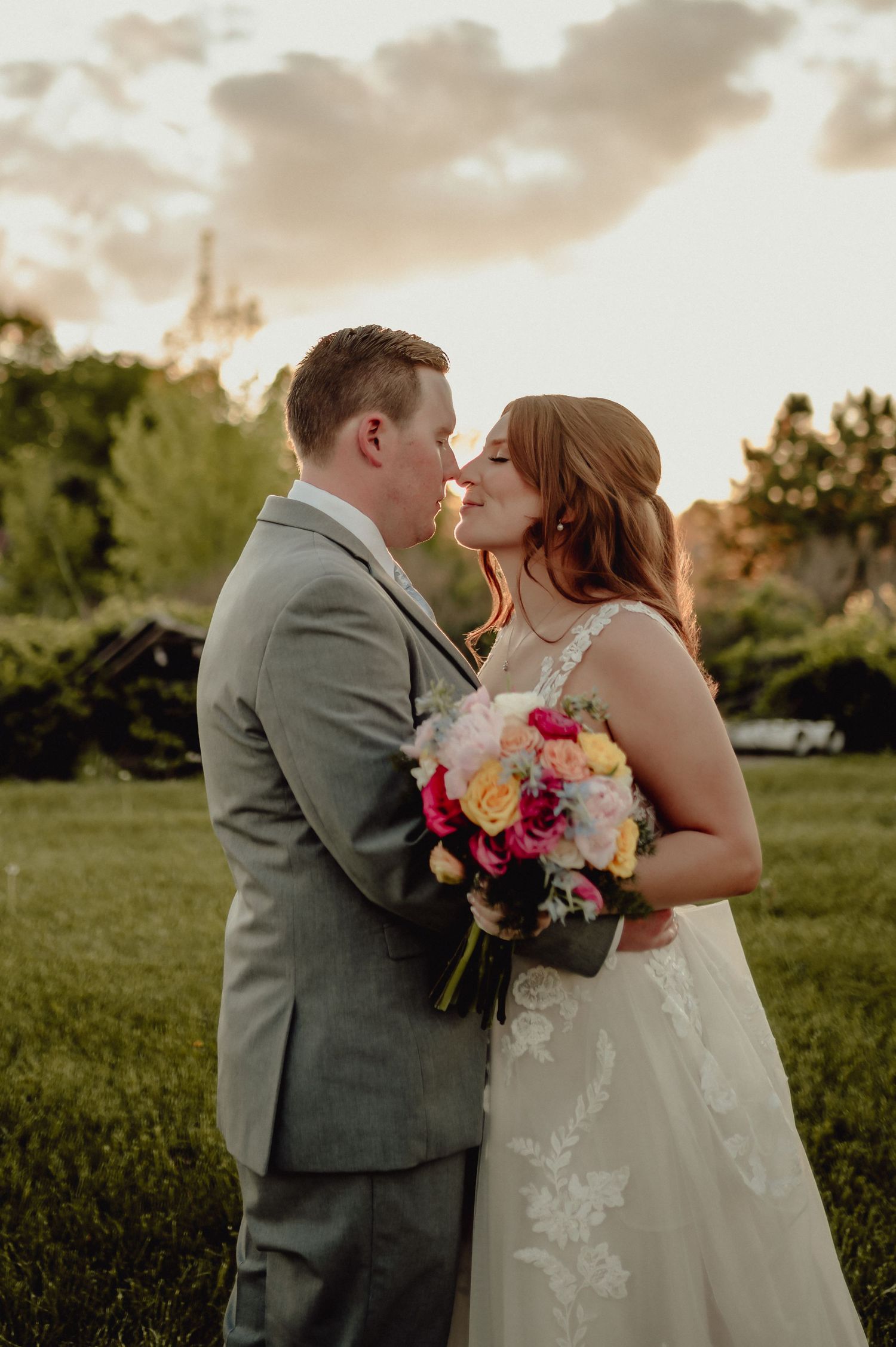 A romantic wedding couple shares a kiss at sunset in a lush green field with colorful bouquet details.