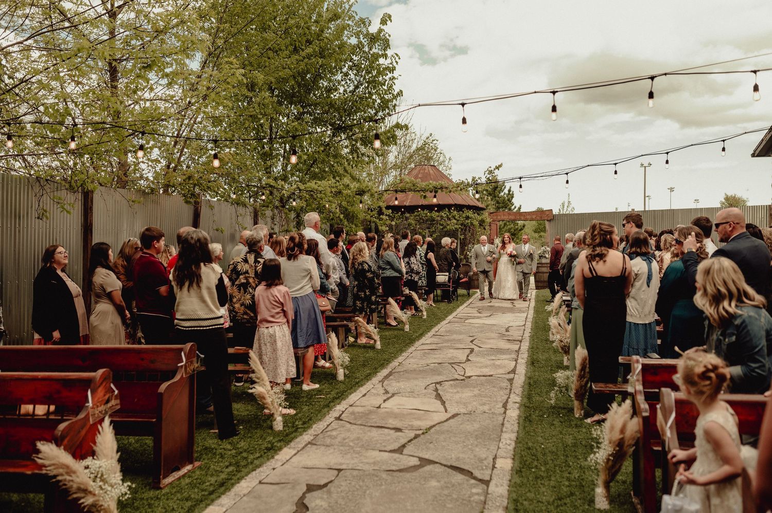 Outdoor wedding reception with string lights and guests gathered in a garden setting during sunset.