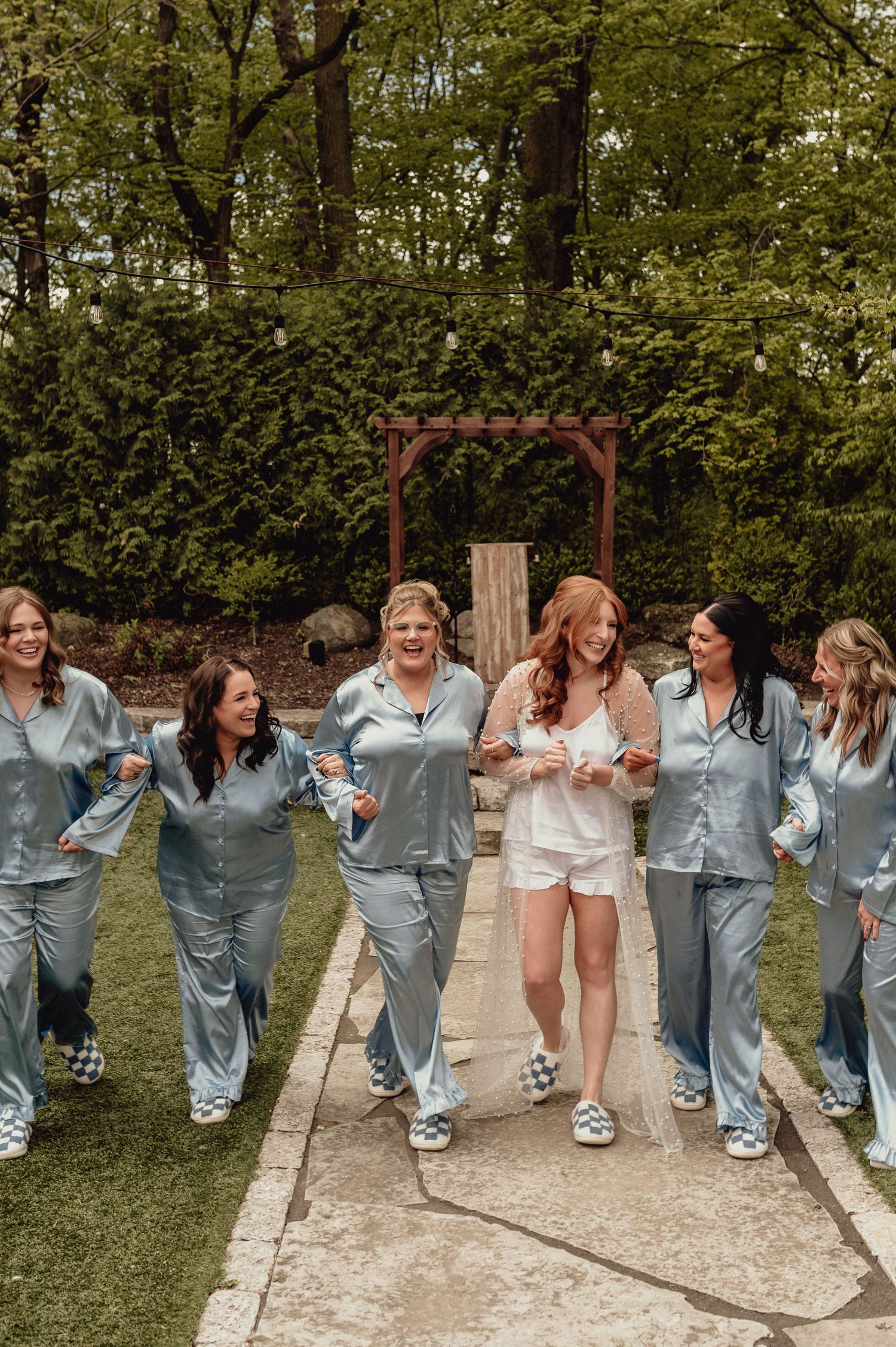 A group of friends in matching gray sleepwear laugh and dance together in a line outdoors during a celebration.