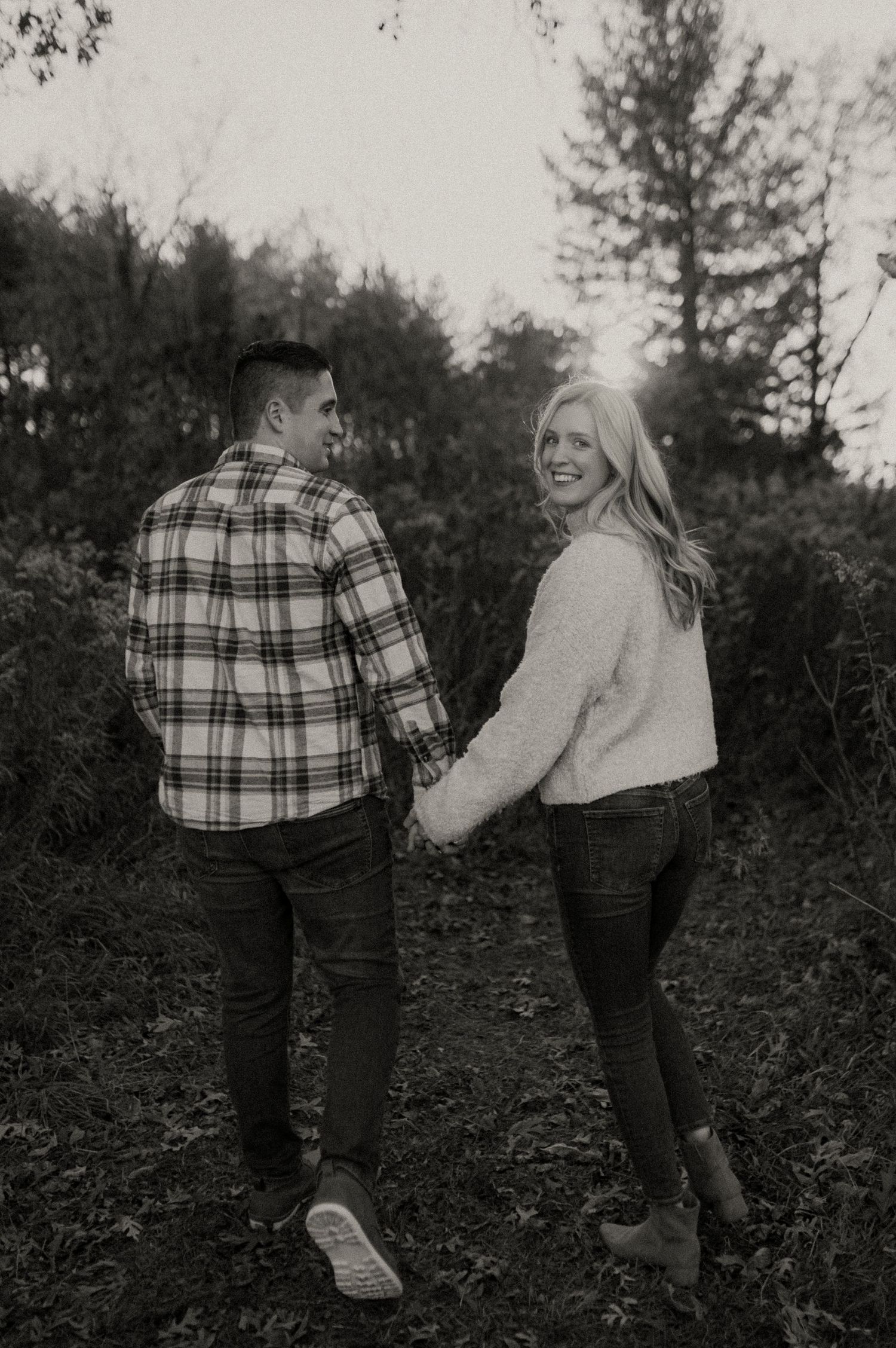 Couple holding hands while walking through wooded area in black and white photo.