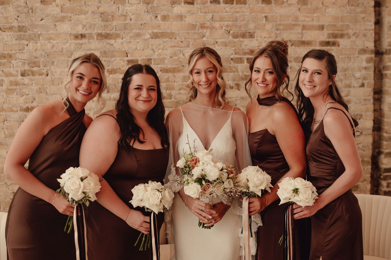 Bridesmaids in brown dresses hold white bouquets while posing against exposed brick wall at wedding celebration.