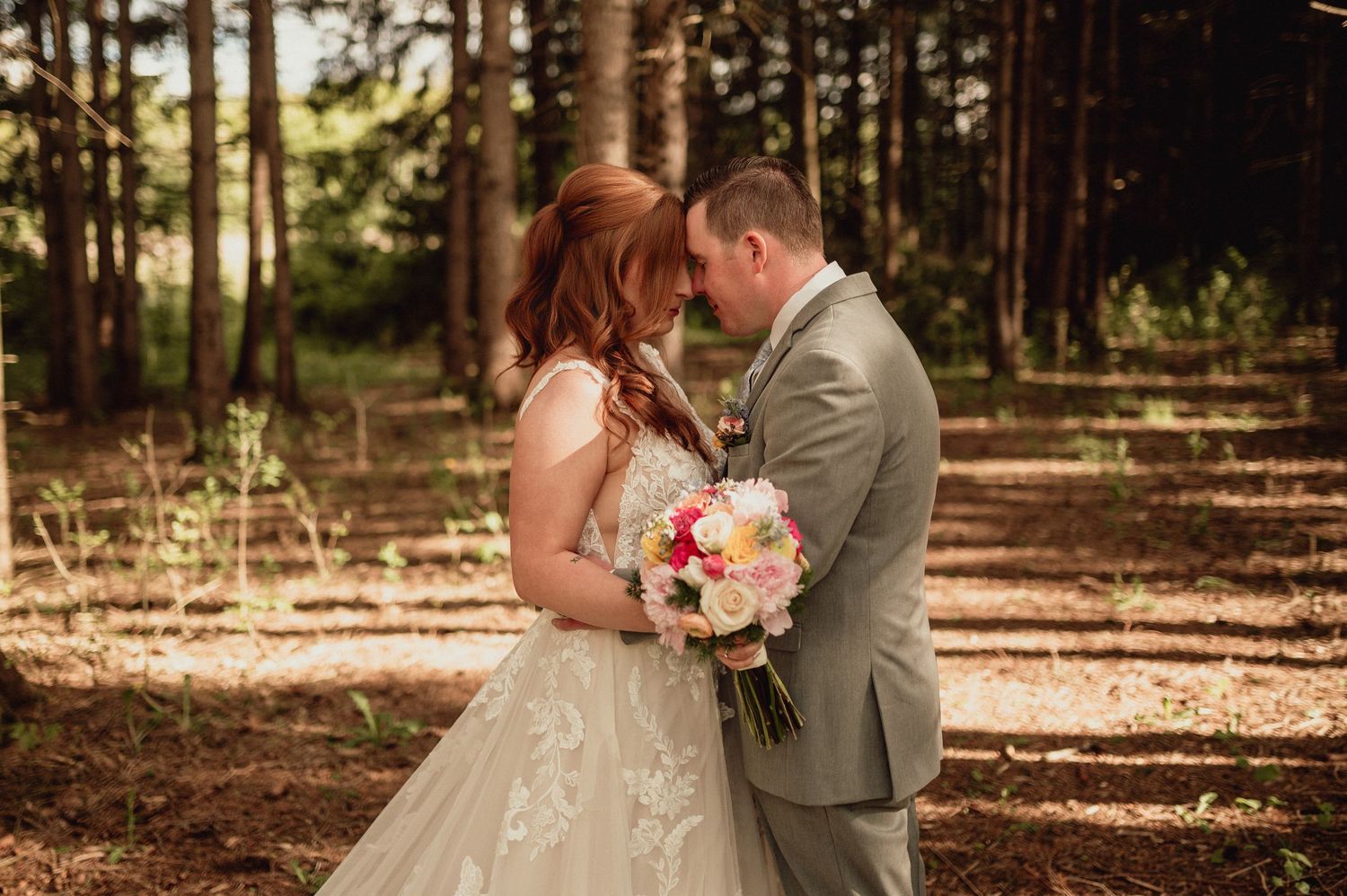 A couple embraces during an outdoor wedding photoshoot in a sunlit forest setting with pine trees in the background.