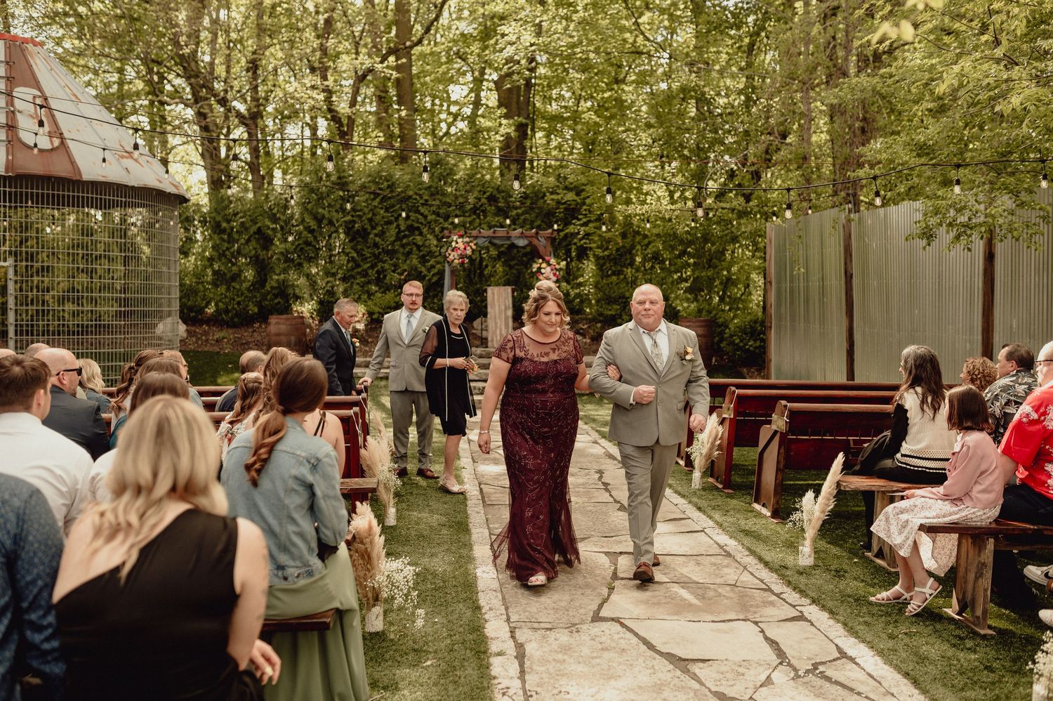 Wedding party members walk down stone pathway during outdoor ceremony while guests look on from seated rows.