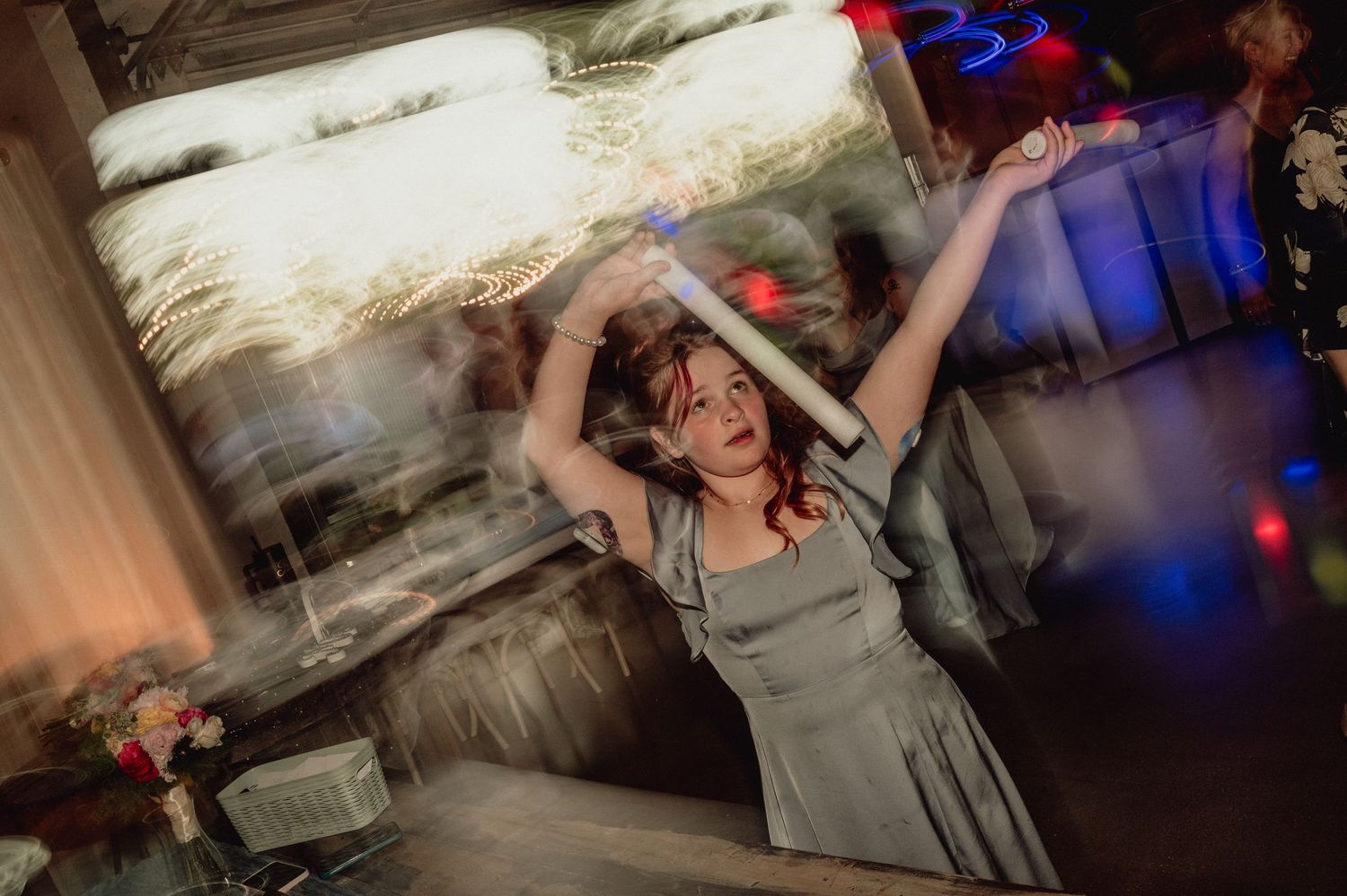 A person in a gray dress dances energetically at a party with colorful lights and decorations in the background.