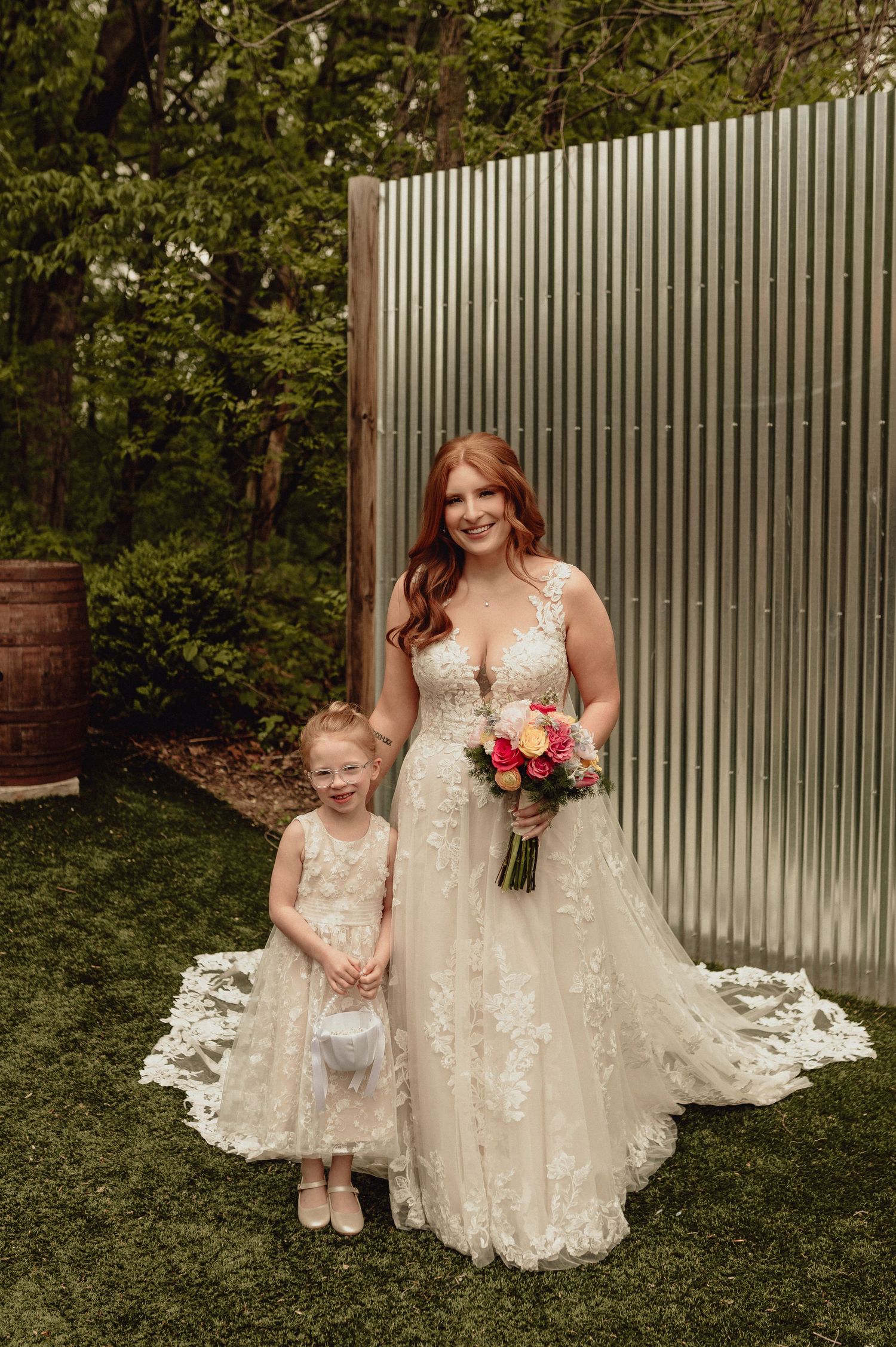 A bride in a lace wedding dress poses with a young child in a matching dress on a grassy lawn near a white fence.