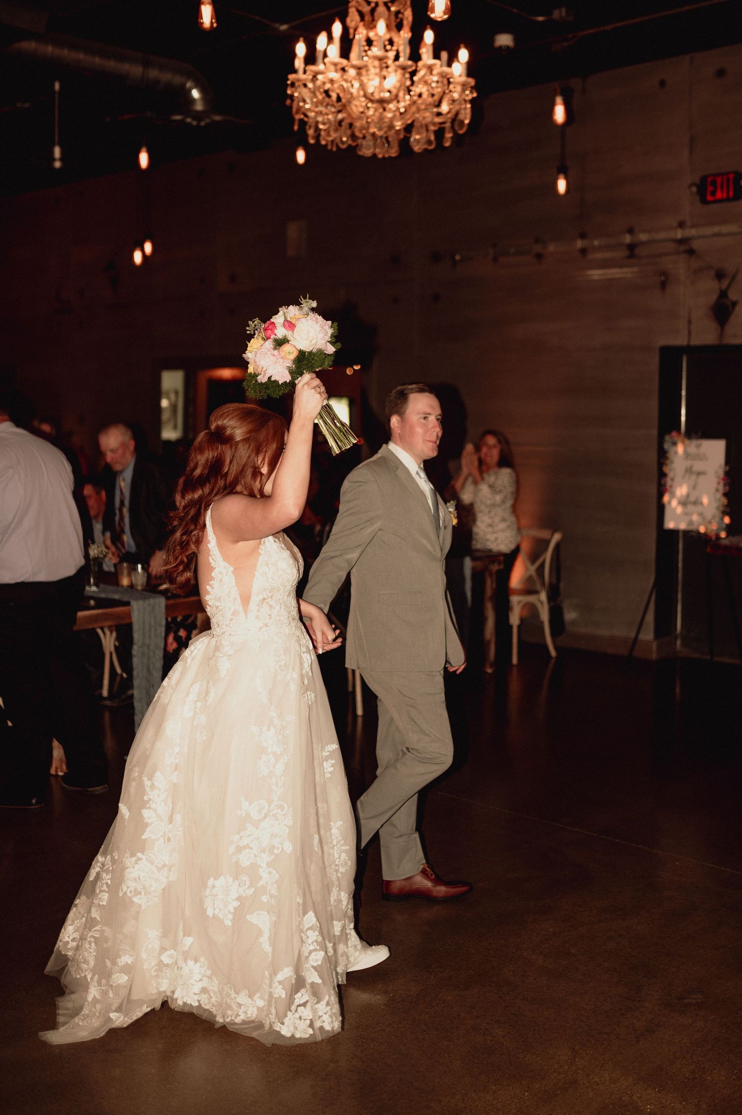 A couple celebrates at their wedding reception under an elegant crystal chandelier in a dimly lit venue.