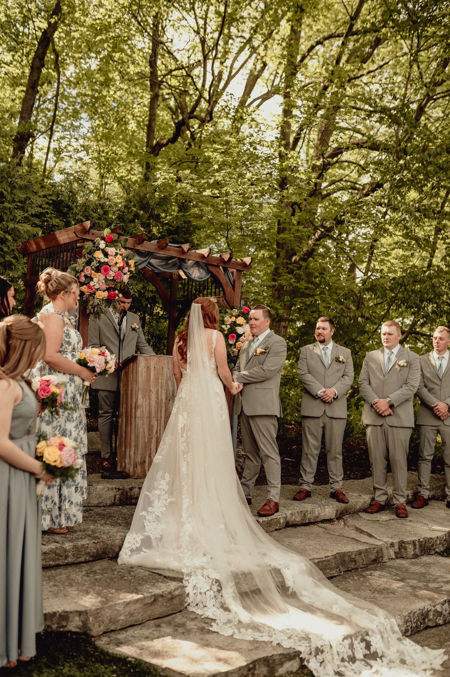 Wedding party in gray attire poses along rustic stone wall in sunlit wooded outdoor setting.