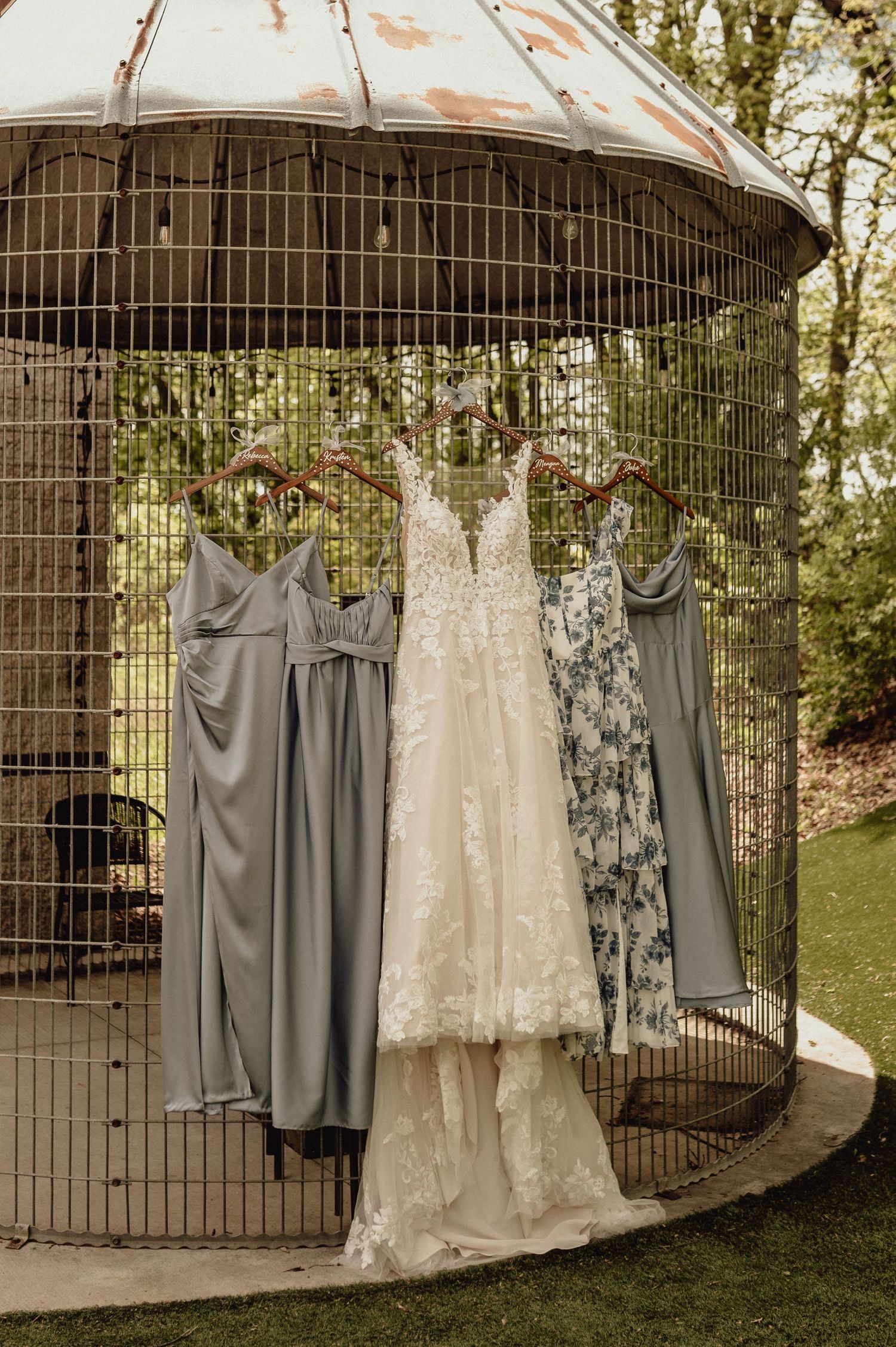 Wedding gown and gray bridesmaid dresses hanging on rustic metal gazebo in outdoor garden setting.