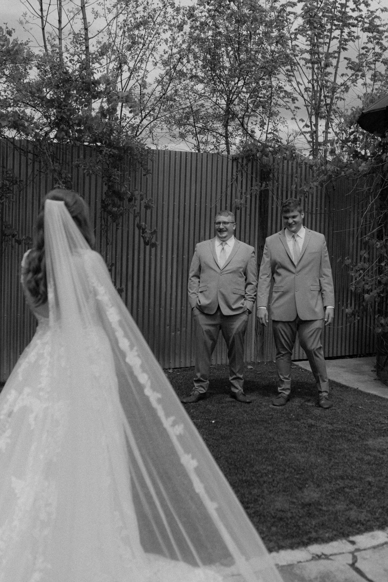 A black and white wedding photo showing a bride in a long veil with two suited men standing in a garden setting.