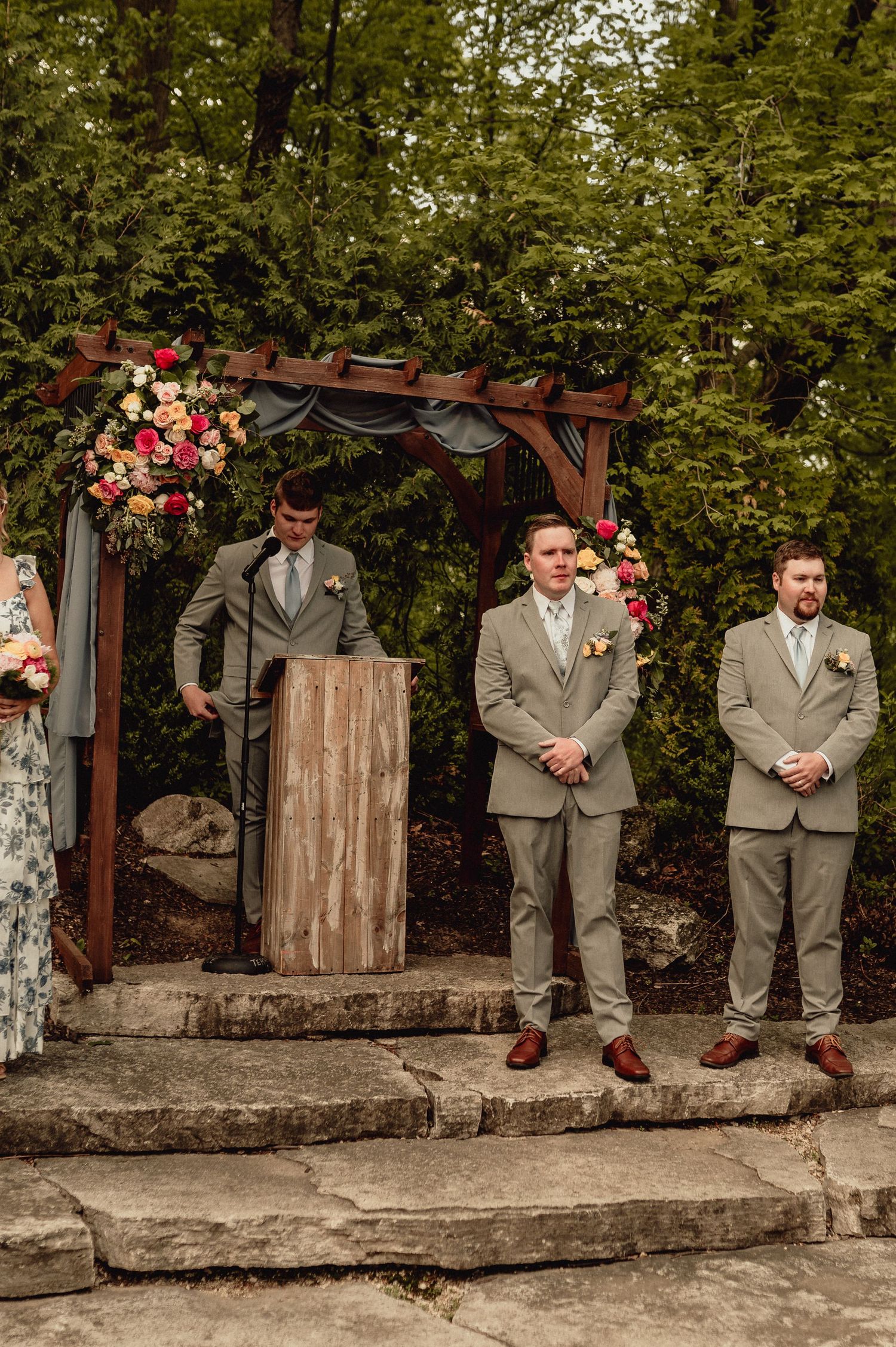 A wedding party in gray suits sits casually on a rustic stone wall with pink floral decorations in the background.