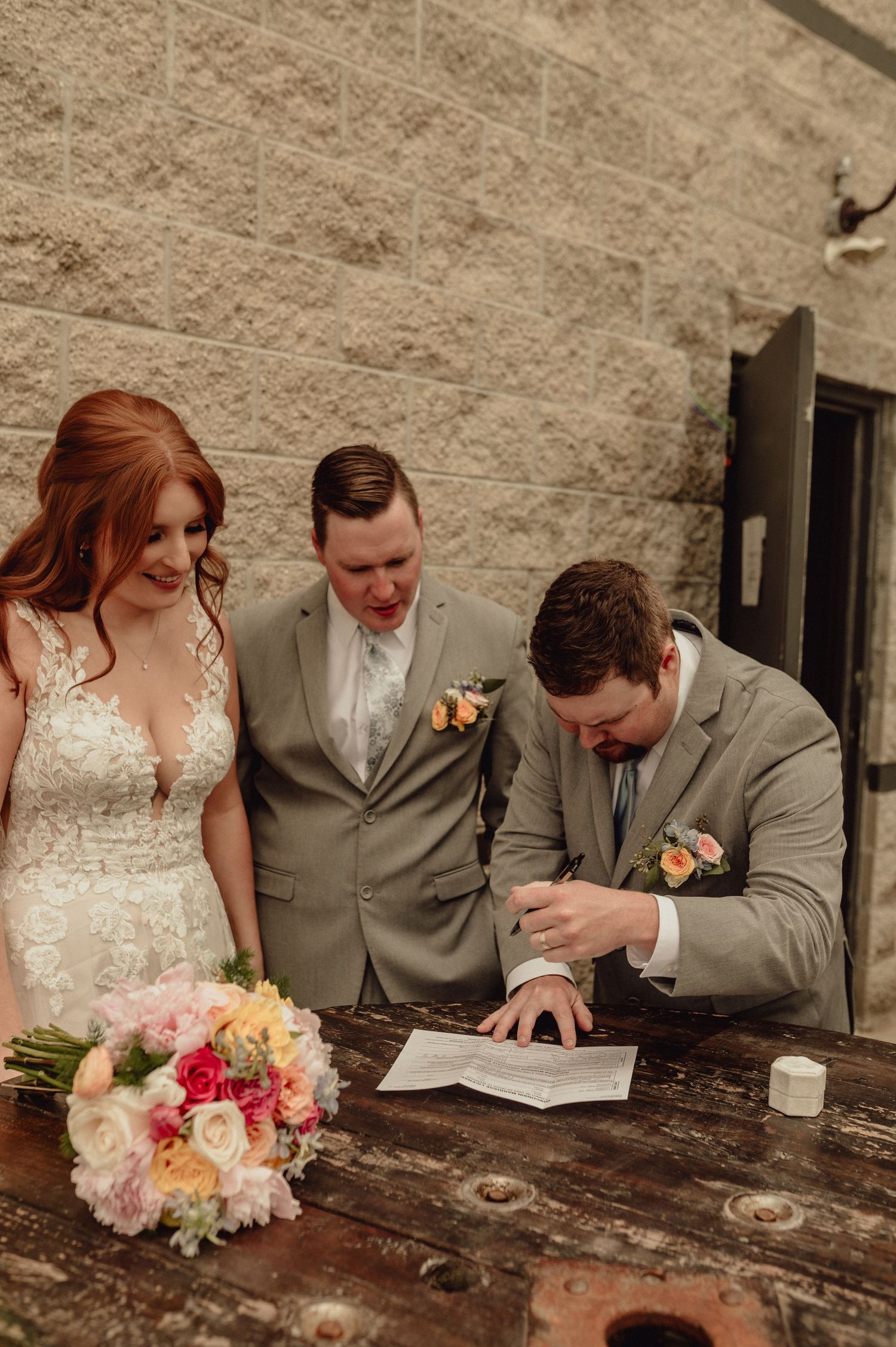 Wedding party signs marriage documents at rustic wooden table with colorful floral bouquet.