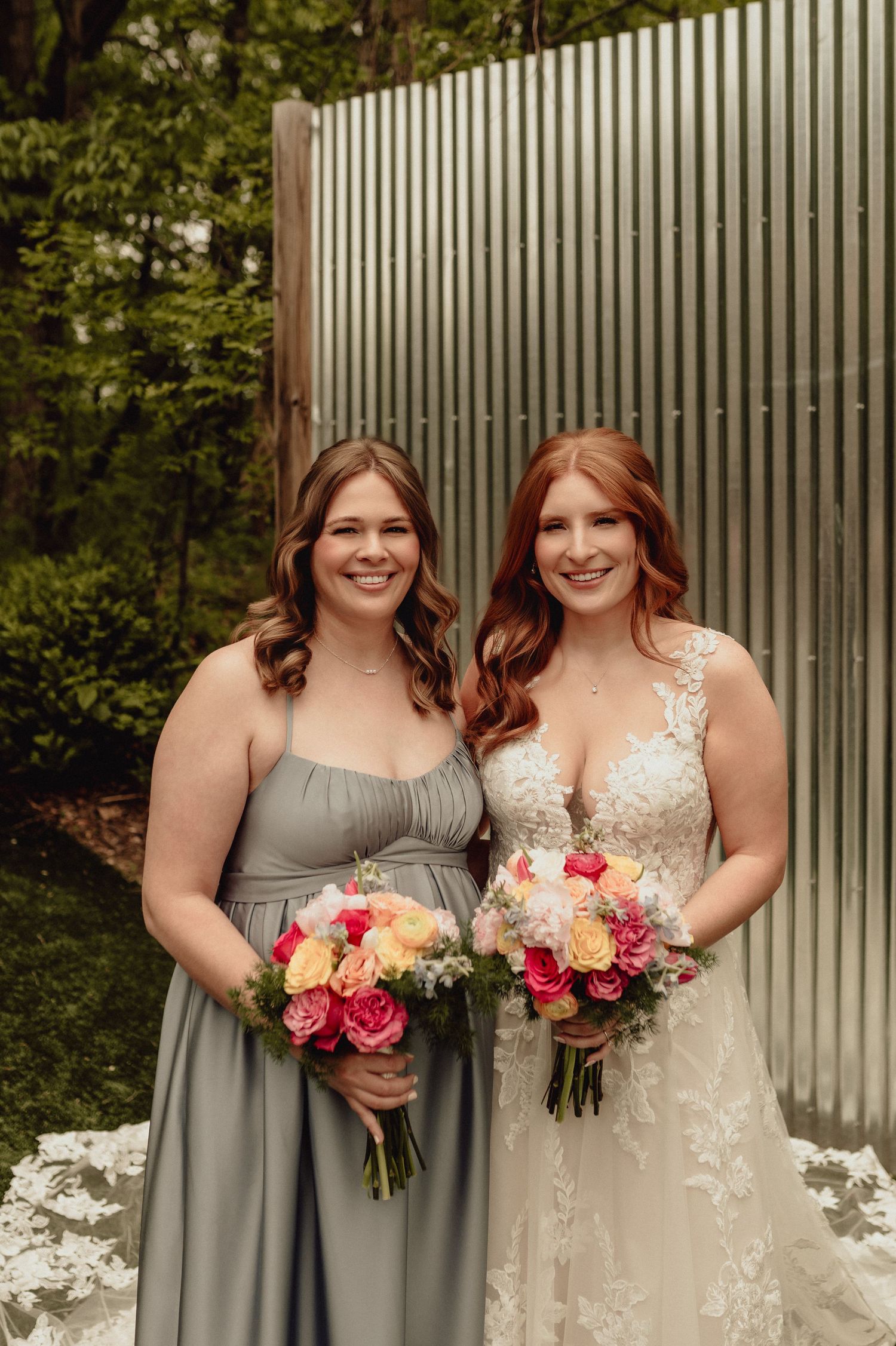 Wedding party members in gray bridesmaid dresses and white bridal gown hold colorful bouquets against metal wall backdrop.